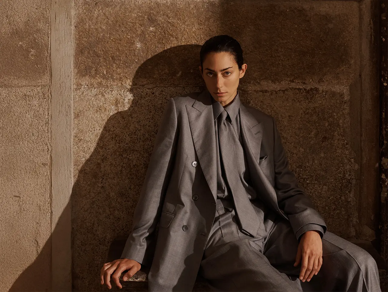 Woman in Brioni gray tailored suit sitting against textured brown wall, looking directly at camera with serious expression.