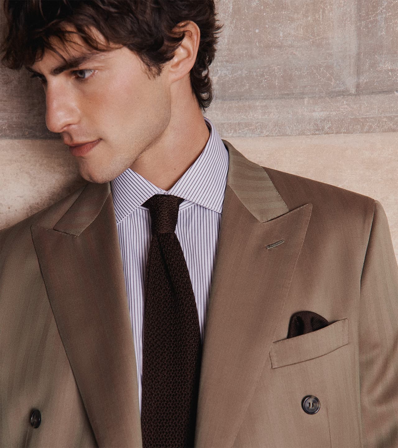 Young man in Brioni double-breasted suit with striped shirt and dark knit tie against textured beige wall background.