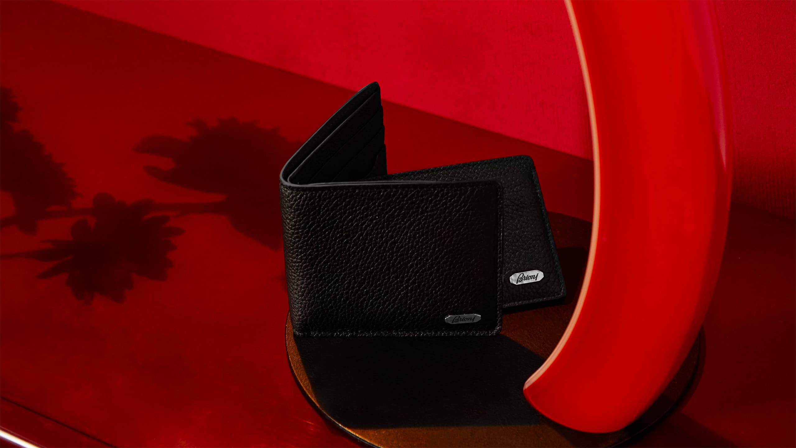 Black leather Brioni wallet displayed on a glossy red surface, with brand logo visible on the exterior.