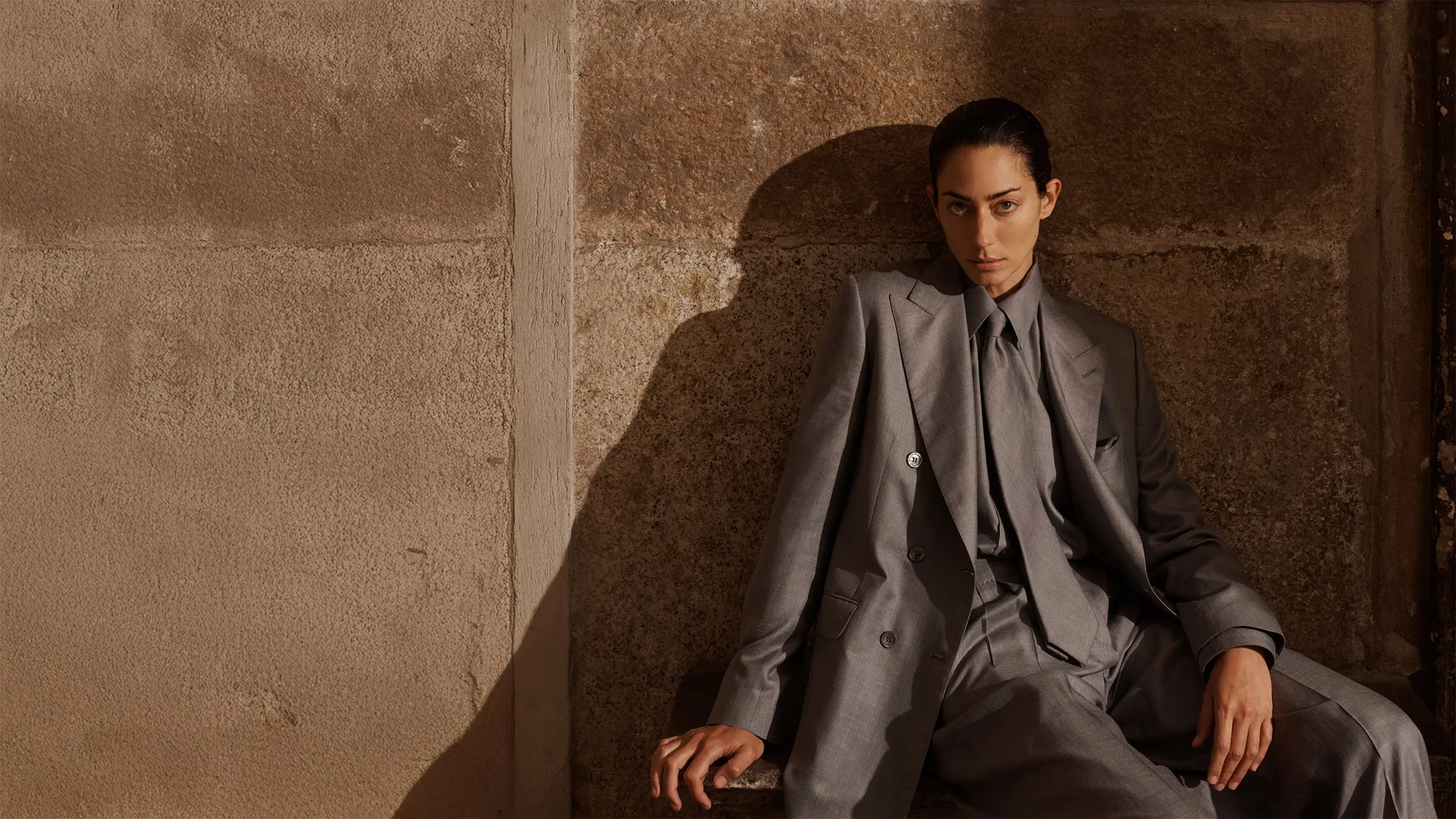 Woman in Brioni gray tailored suit sitting against textured beige wall with dramatic shadow, looking directly at camera.