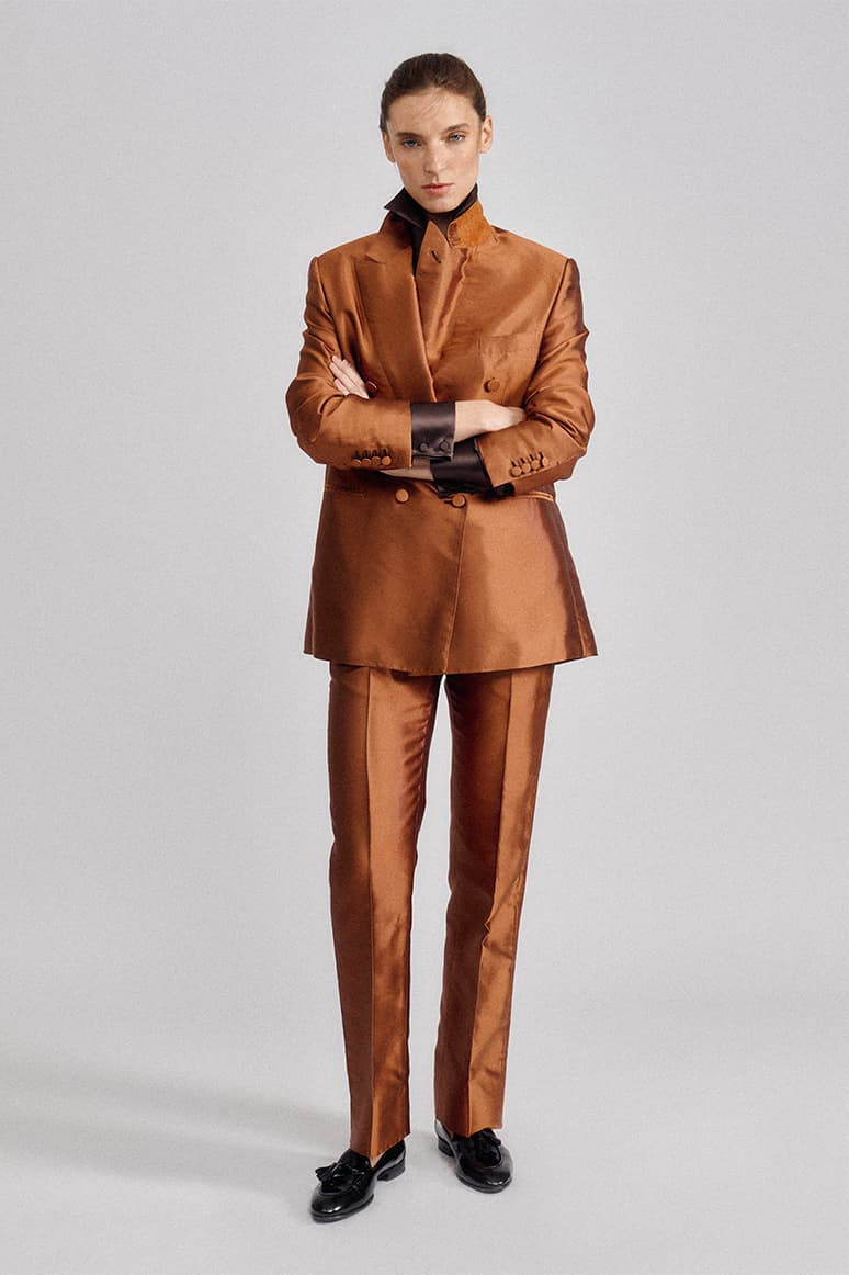 Woman in Brioni copper-colored satin suit with black collar and black loafers, arms crossed, standing against gray background.