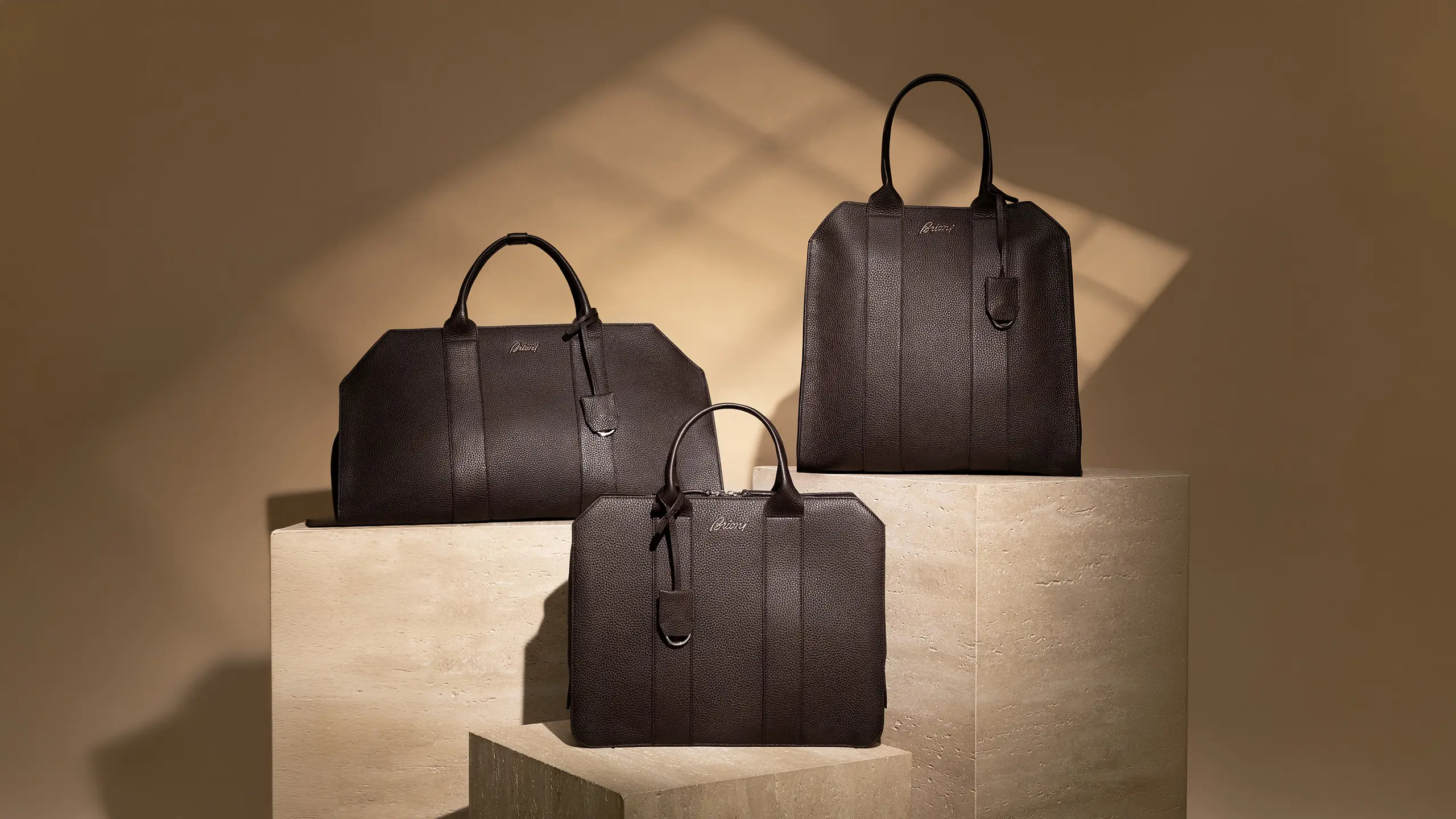 Three dark Brioni brown leather handbags displayed on beige stone pedestals against a tan wall with geometric shadow patterns.