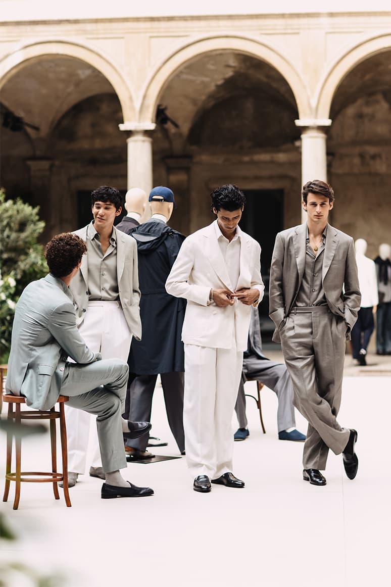 Models in elegant Brioni's SS26 collection suits posing in a historic courtyard with arched columns.