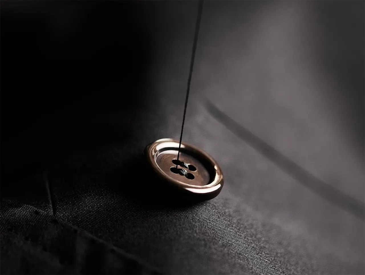 Close-up of a gold button with black thread on dark Brioni fabric, creating a subtle shadow in dramatic lighting.