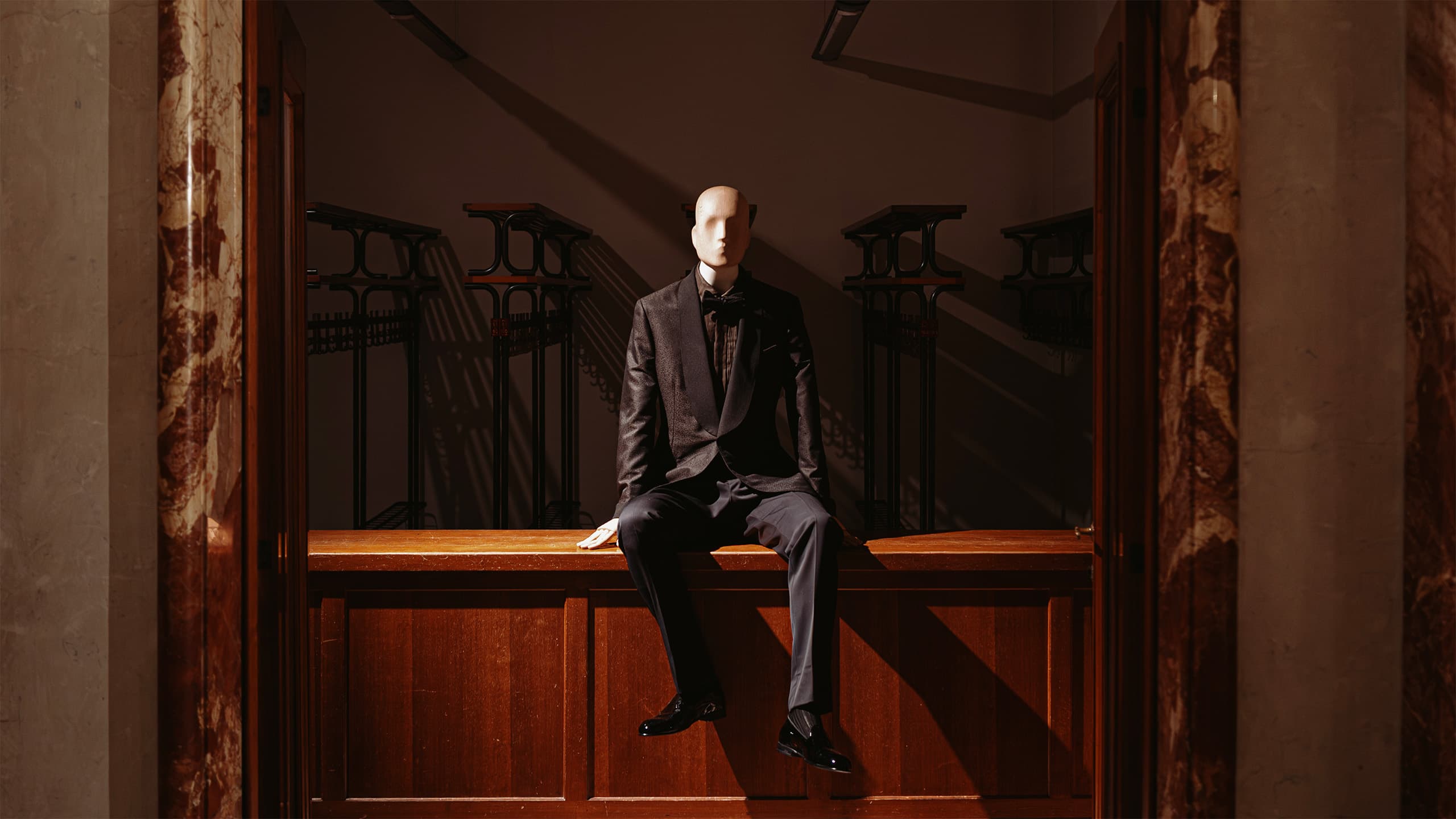 Mannequin in Brioni dark suit sitting on wooden bench in elegant hallway with marble columns and ornate railings.