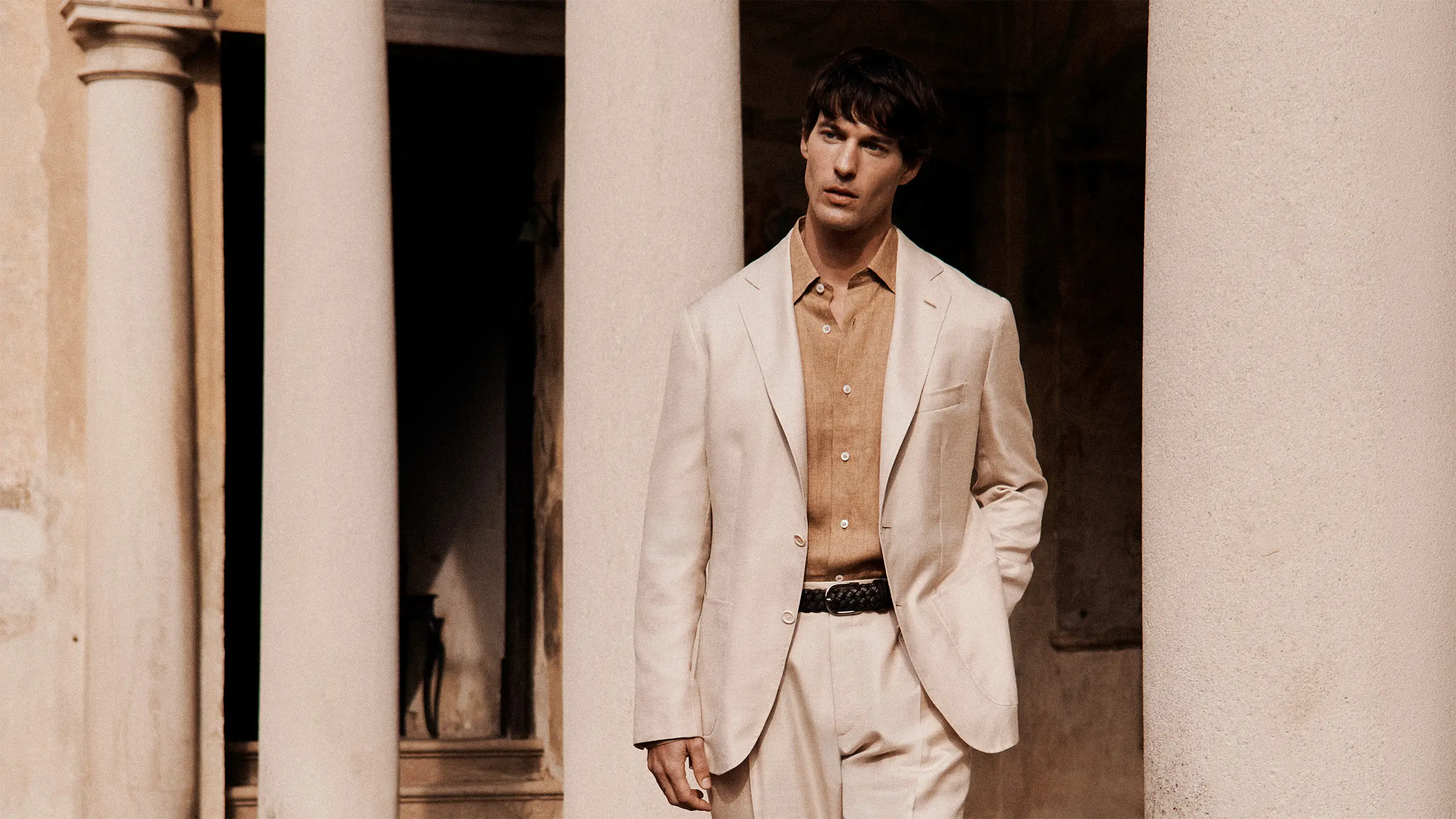 Man in beige Brioni suit and tan shirt standing between white columns in warm-toned architectural setting with dark doorway behind.