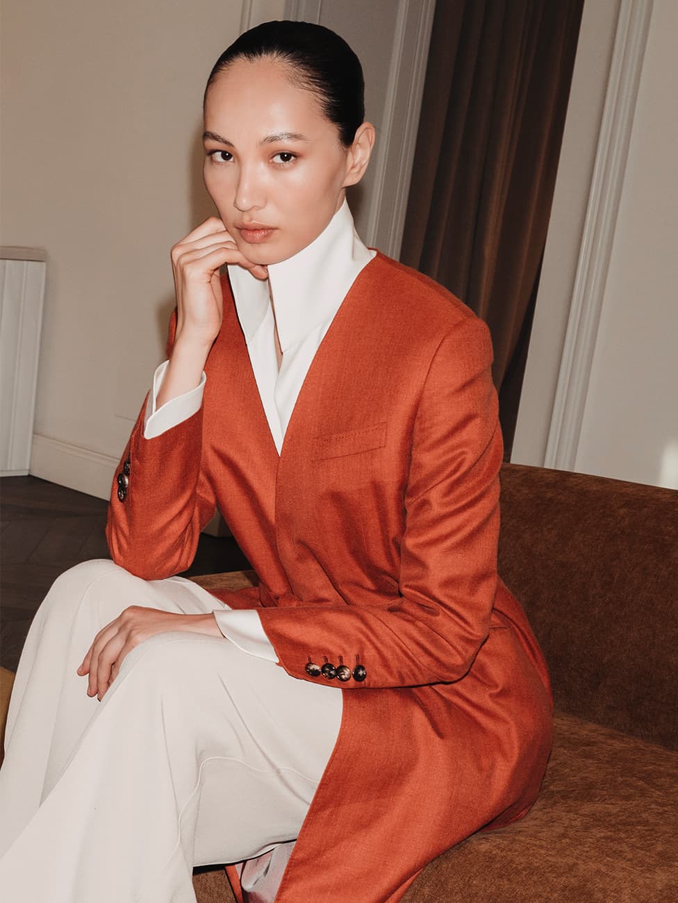 Woman with slicked-back hair wearing an orange Brioni blazer over white collared shirt and white pants, seated indoors.