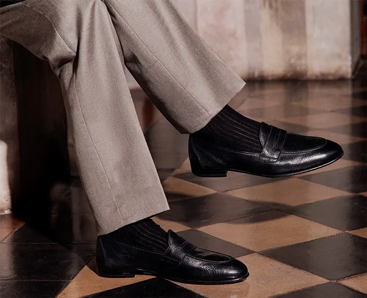 Person wearing beige trousers and black leather loafers standing on a checkered brown and tan floor.