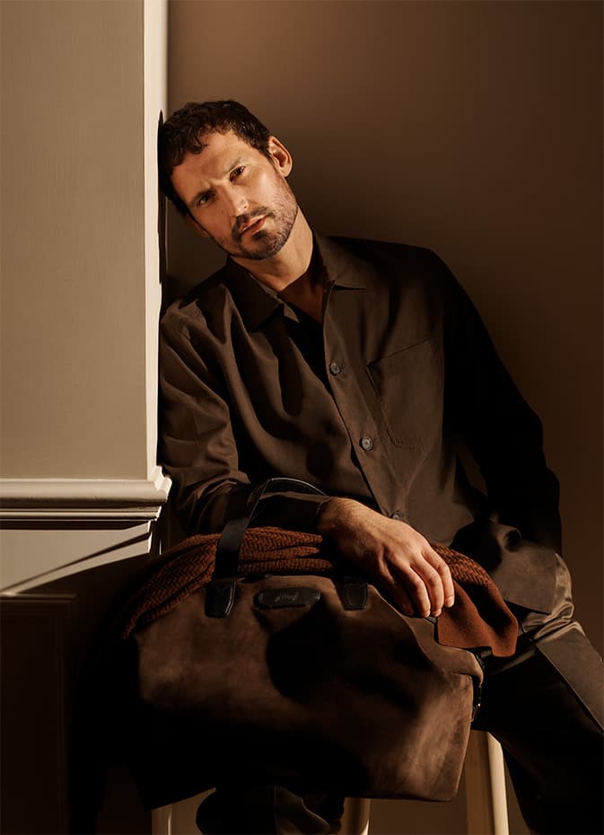 Man in brown Brioni shirt sitting by window with warm lighting, holding a brown bag with rope handles on his lap.
