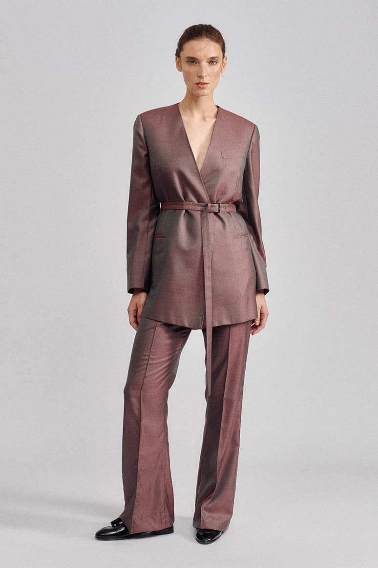 Woman wearing a Brioni mauve belted wrap blazer and matching wide-leg trousers with black shoes on a gray background.