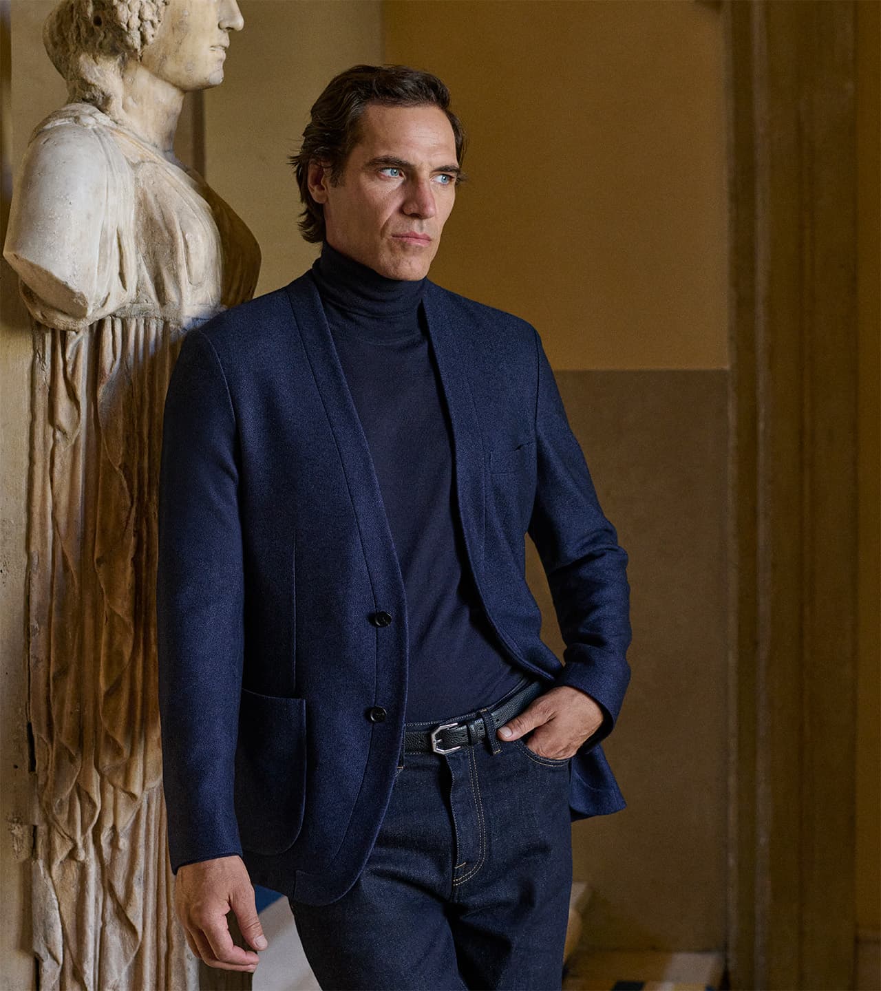 Man in Brioni navy blazer and turtleneck standing beside classical stone statue in warm-toned interior setting.