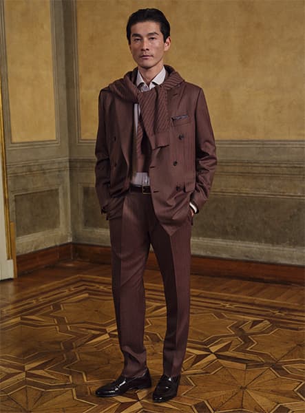 Model in elegant brown brioni suit on patterned wooden floor against gold-toned wall.