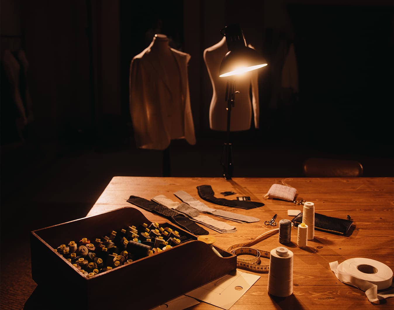 Tailor's workspace with mannequins, thread spools, fabric swatches, and measuring tape illuminated by warm lamp light.
