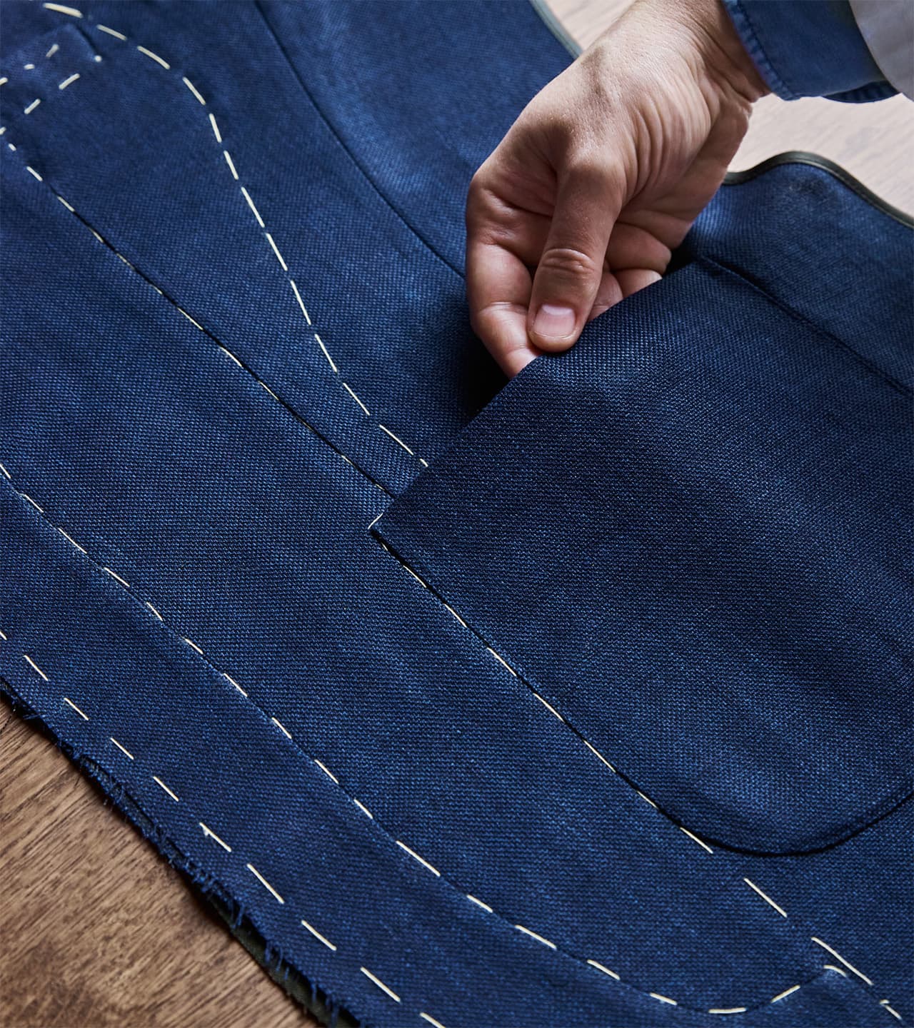 Hand placing a navy blue Brioni suit jacket pocket with white basting stitches on matching fabric during tailoring process.