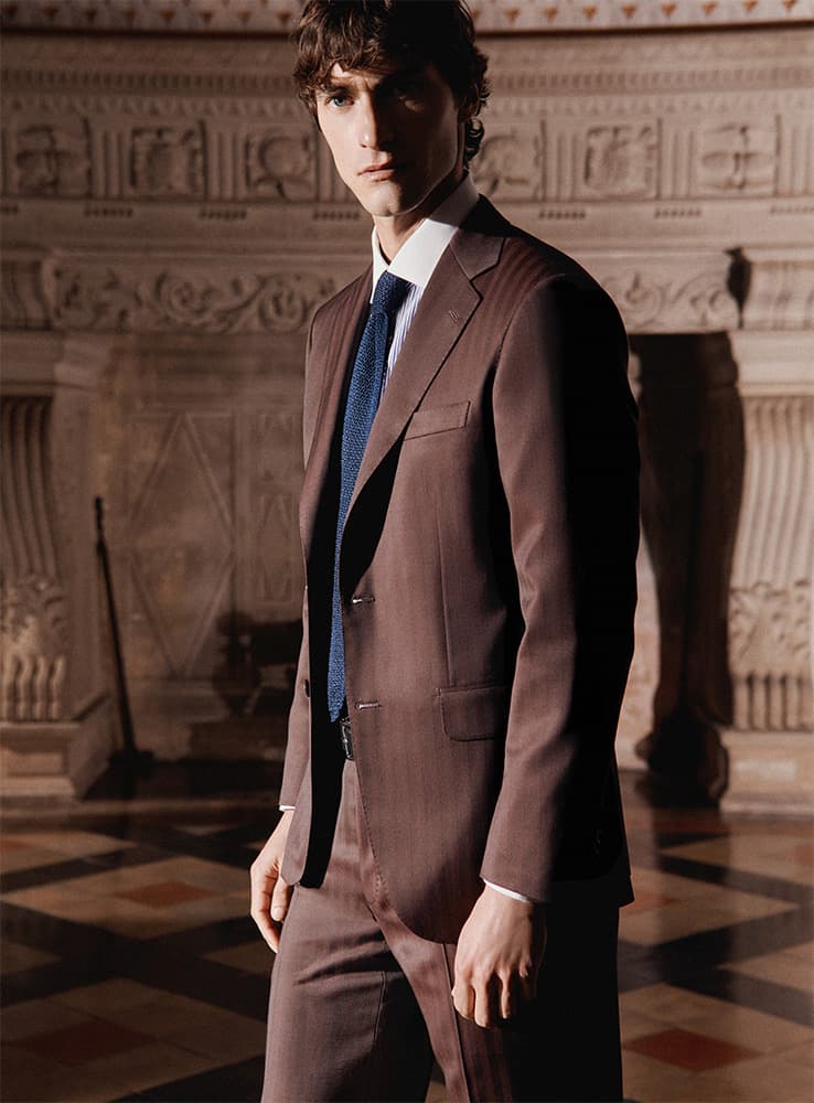 Man in Brioni brown Iride suit with blue tie standing in elegant room with ornate stone walls and decorative architectural details.