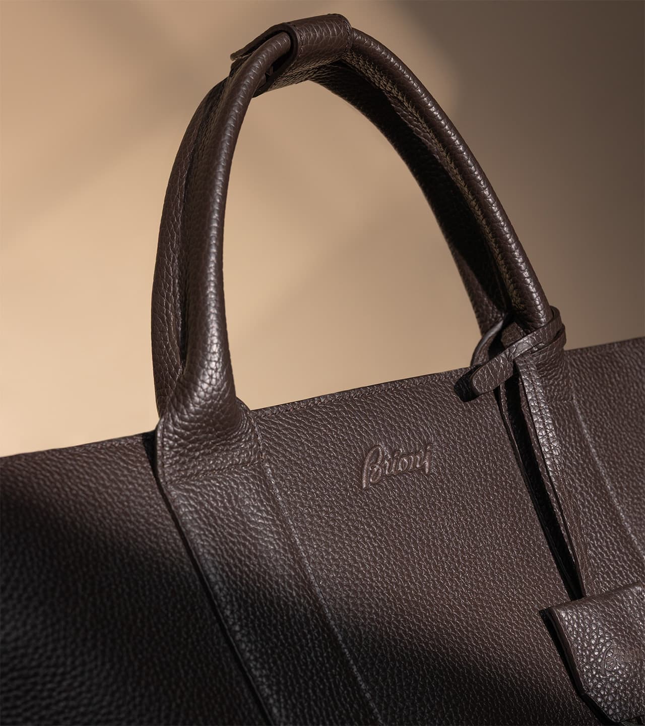 Close-up of a brown Brioni leather handbag with textured surface, knotted handles, and embossed Brioni logo against a beige background.