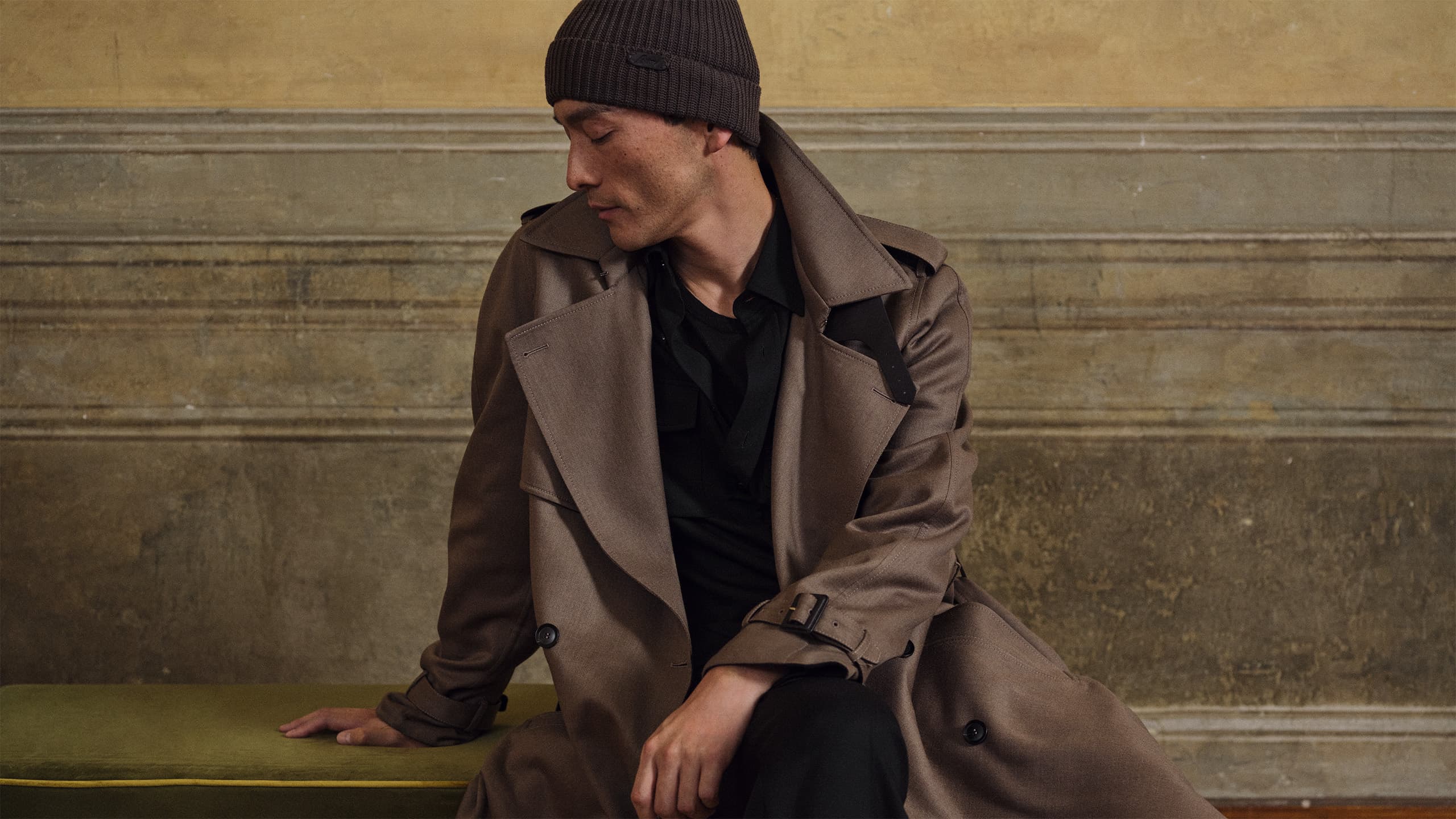 Person in Brioni brown trench coat and black beanie sitting thoughtfully against beige wall background.
