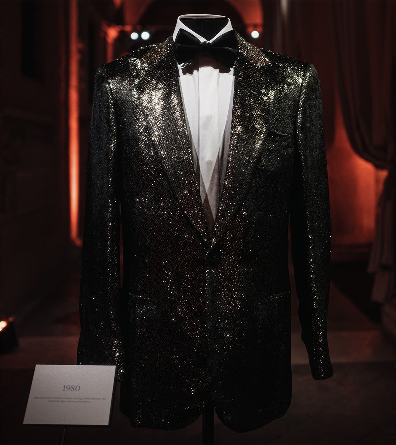 Brioni's sparkly black tuxedo jacket with white shirt and bow tie on display, dated 1980, against a dark red background.