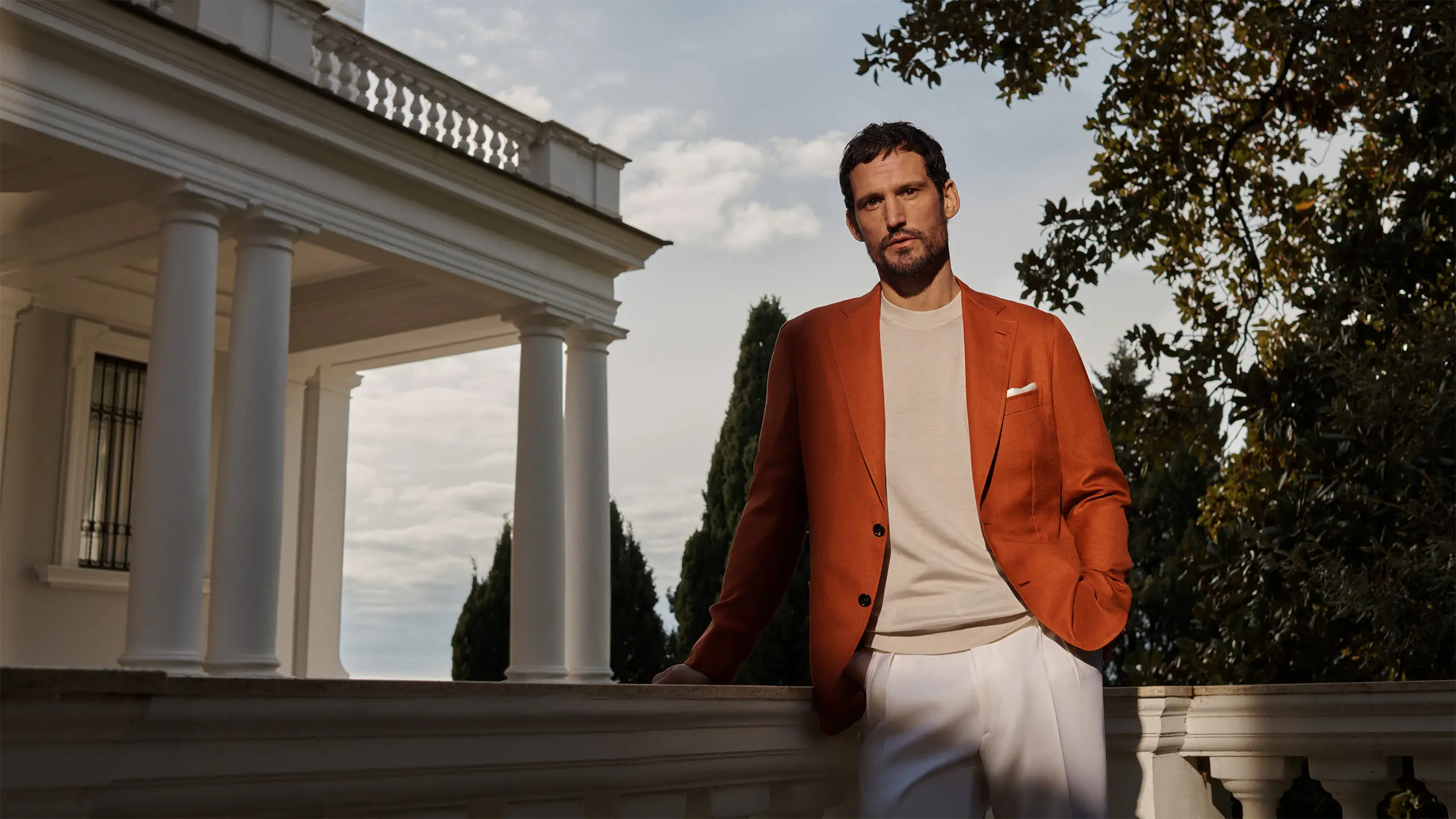 Man in Brioni brick Soffio blazer and cream shirt stands outdoors near white columned building with trees in background under cloudy sky.
