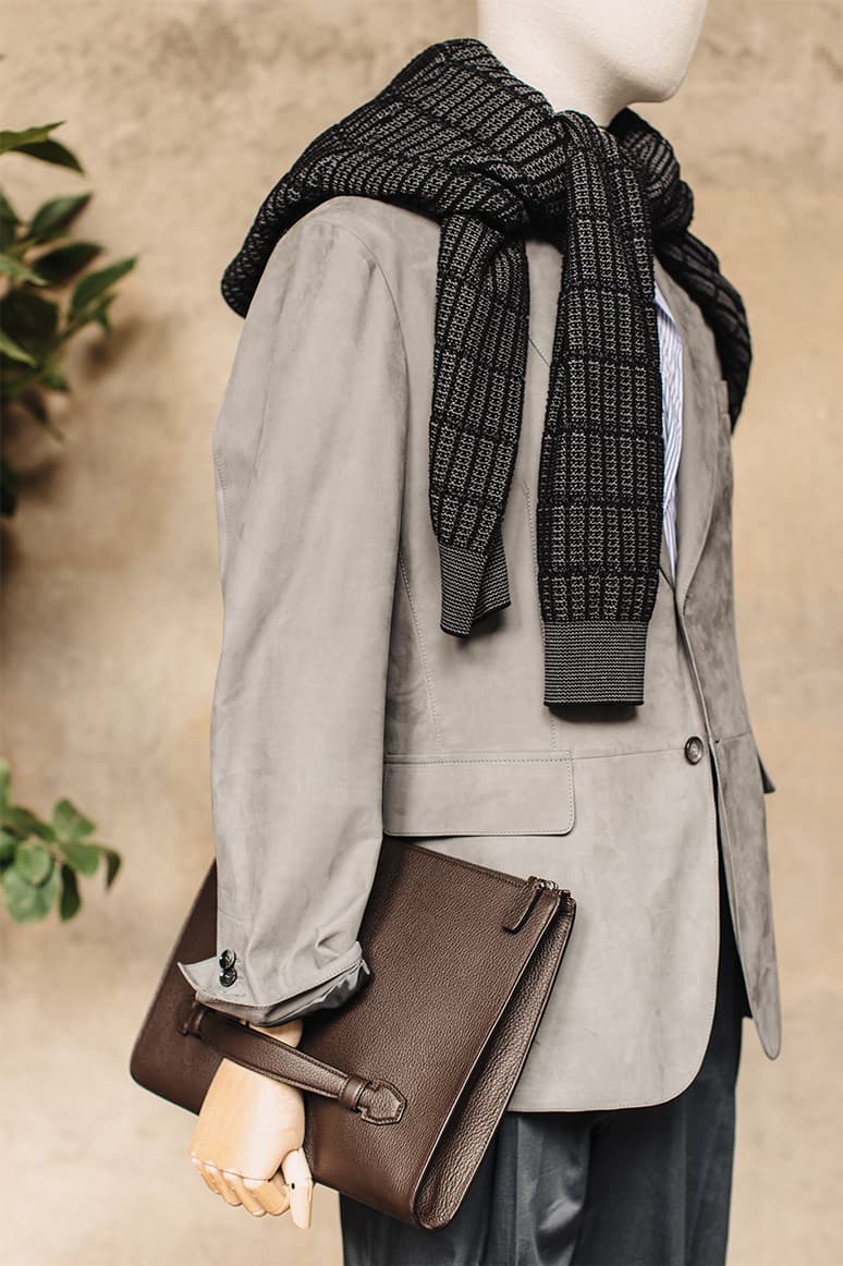 Mannequin wearing Brioni's light gray blazer with black patterned scarf and holding brown leather clutch against beige background.
