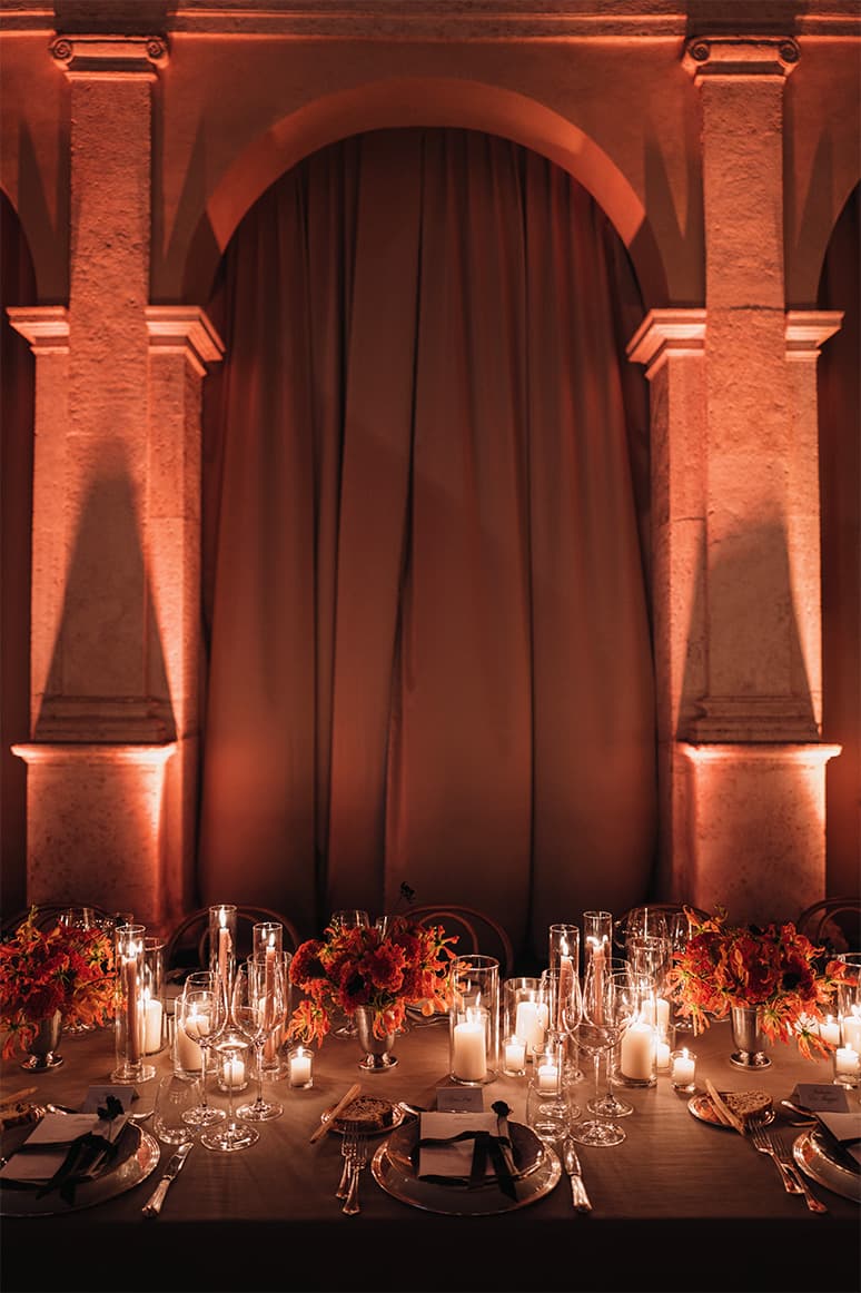 Elegant dining table with candles and orange flowers against dramatic stone archway illuminated in warm amber light.