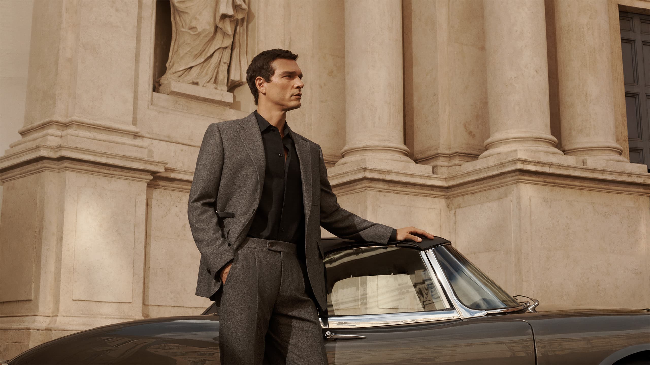 Man in Brioni grey jacket standing beside vintage car against classical stone building with columns and statue.