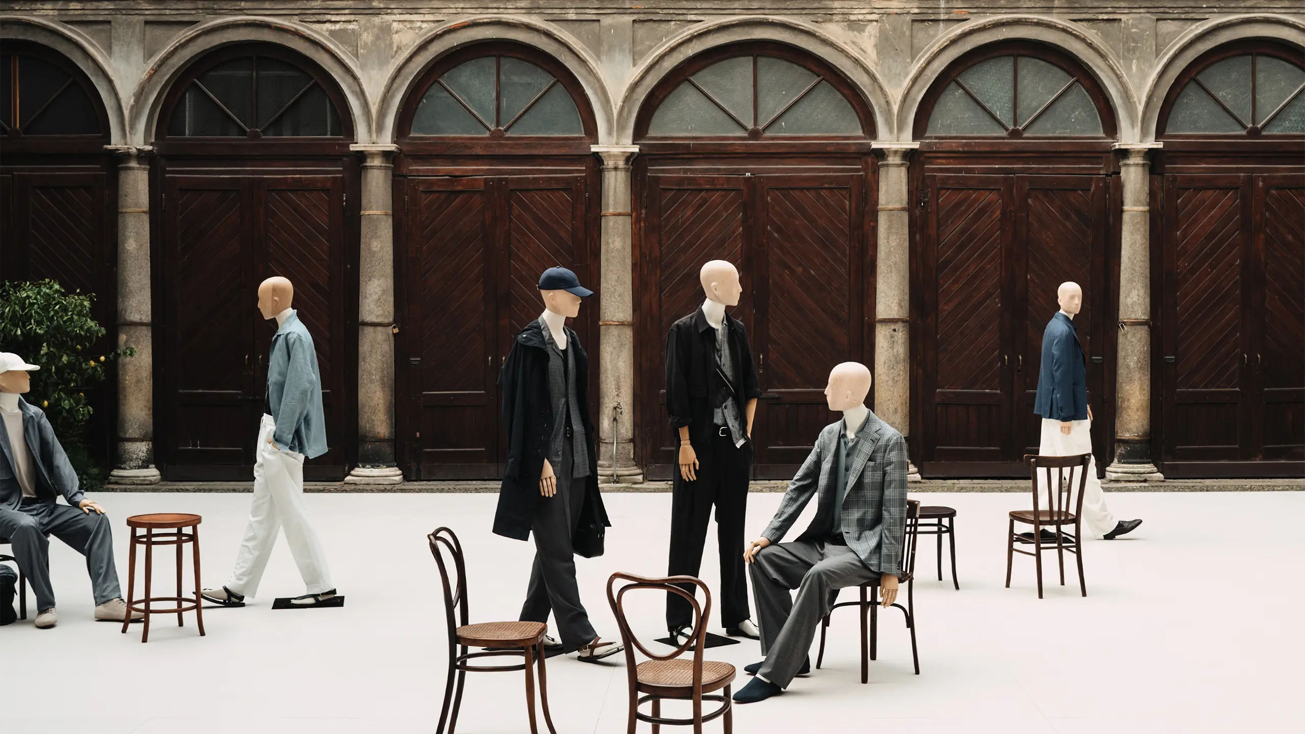 Mannequins in Brioni formal attire arranged with wooden chairs in a courtyard of a historic building with arched doorways.