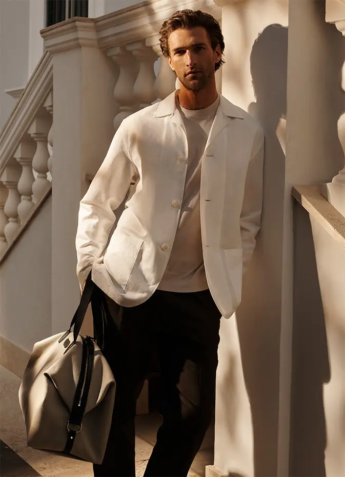 Man in Brioni white blazer and black pants holding beige bag stands by white columns in warm sunlight.
