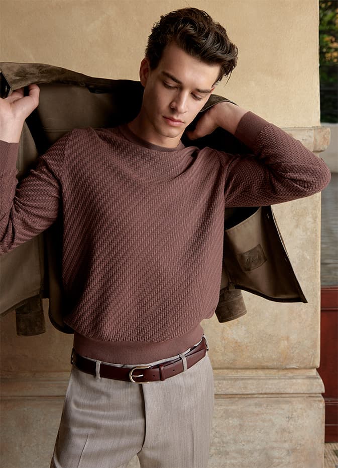Model in Trama burgundy textured sweater removing tan jacket, wearing light trousers with brown belt against beige wall.