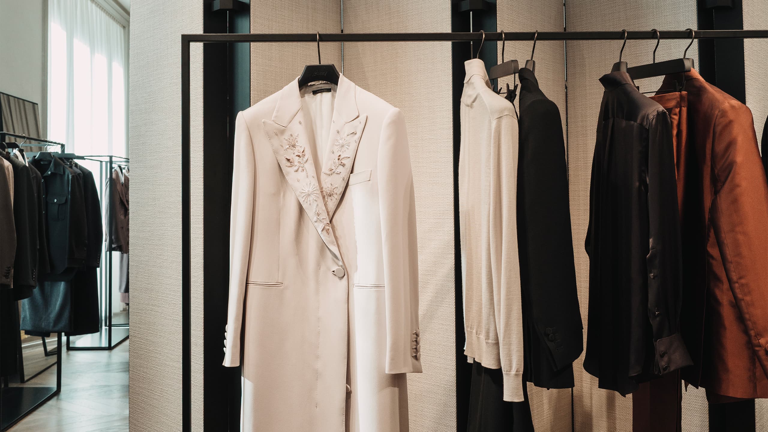 Modern clothing Brioni boutique displaying white embroidered blazer on black rack with neutral and rust-colored garments in background.