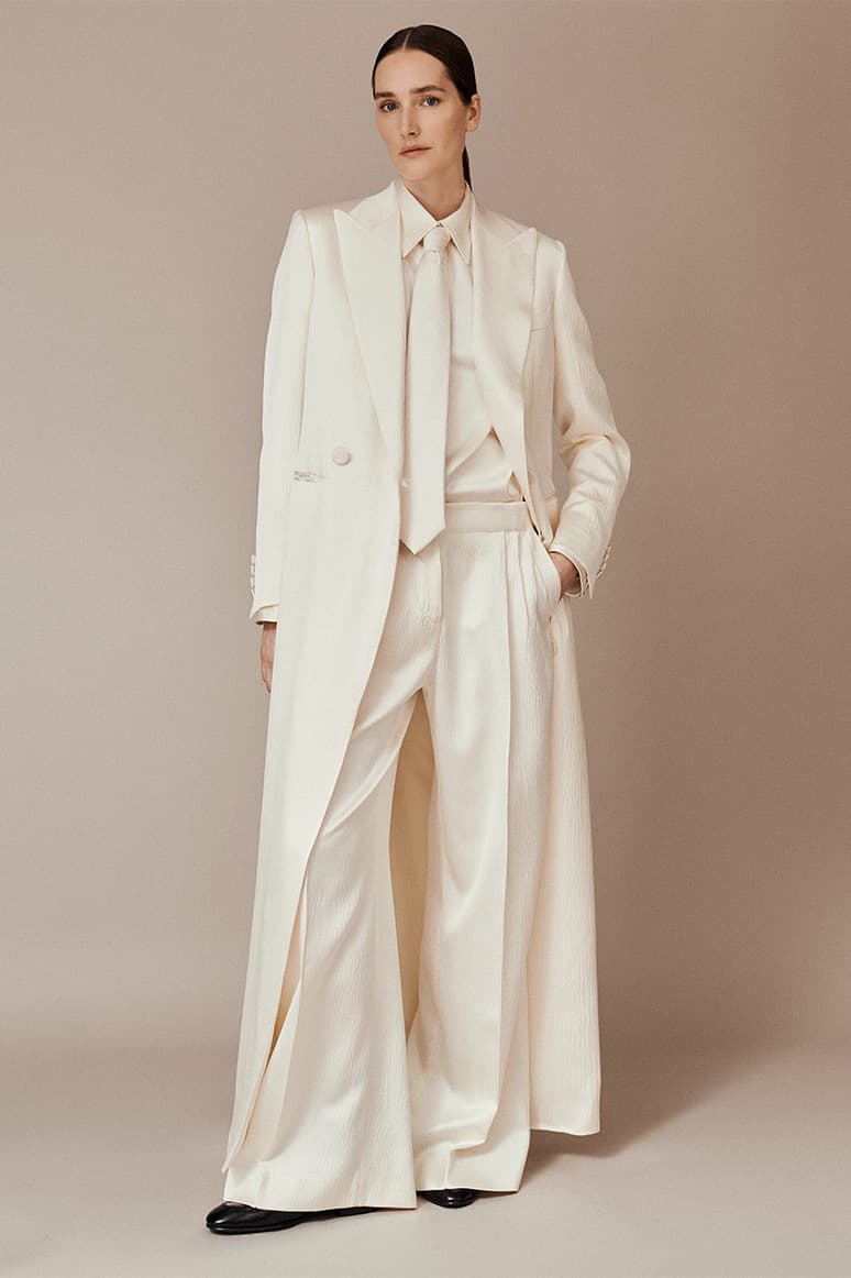 Woman in elegant Brioni cream-colored suit with long coat, wide-leg trousers, and collared shirt against beige background.