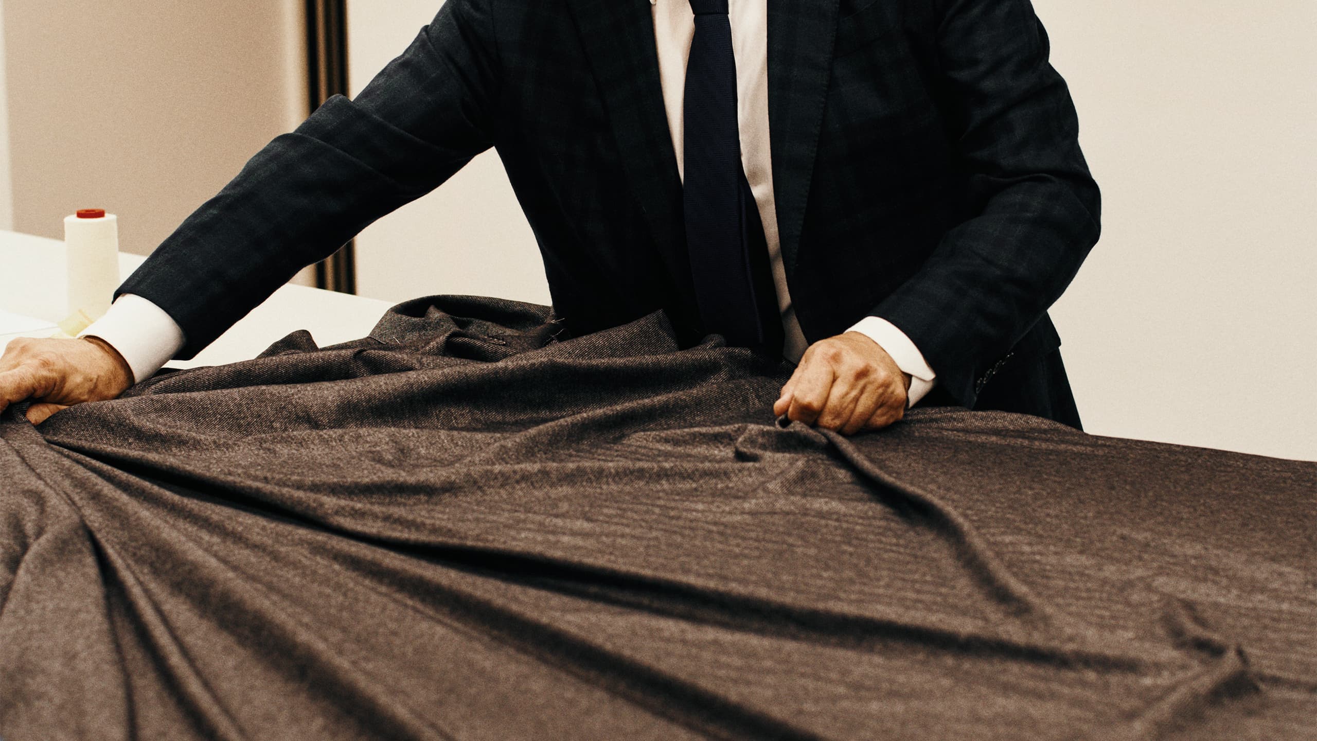 A tailor in black suit examining brown fabric material on a table, with hands carefully arranging the textile.