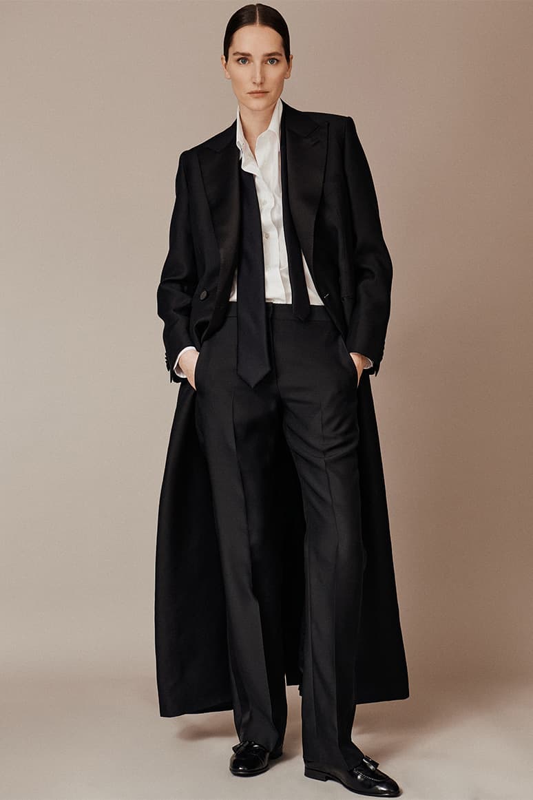 Woman in Brioni black tuxedo with long coat and white shirt standing against beige background in formal outfit.