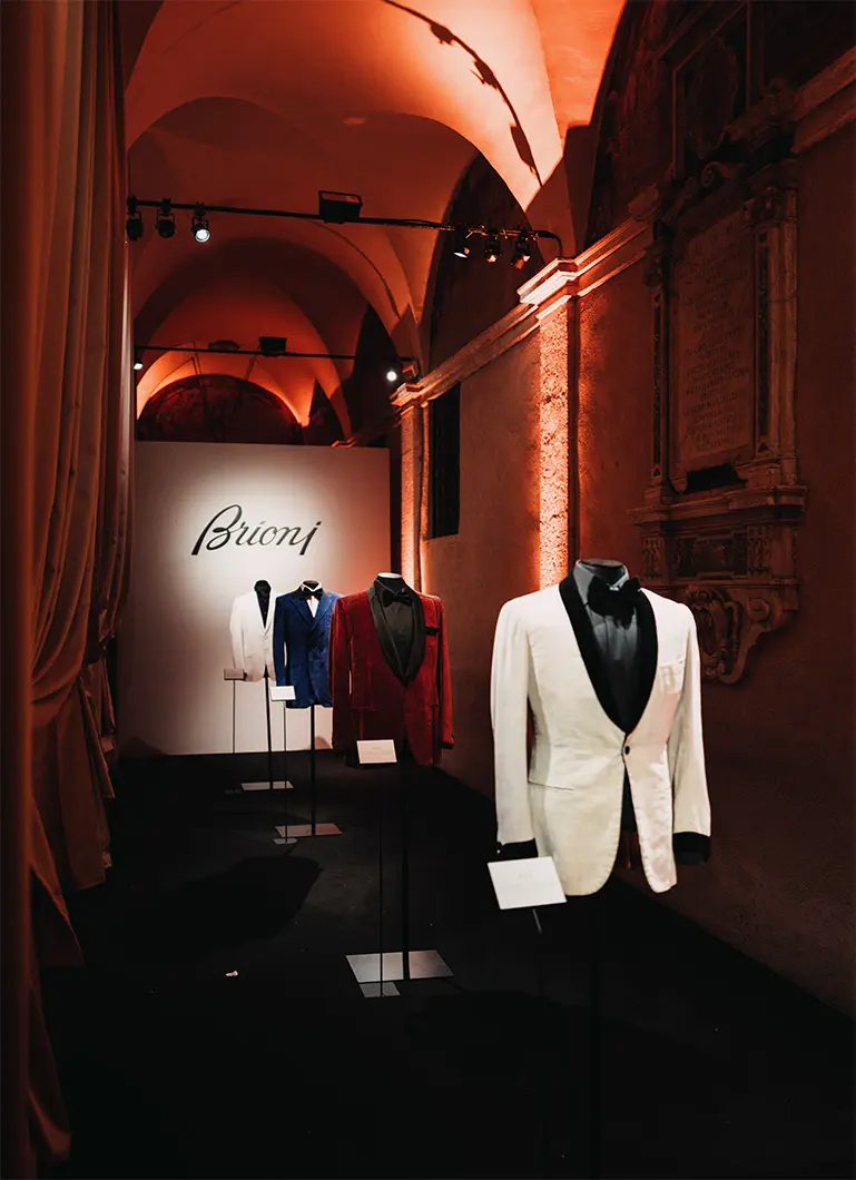 Elegant Brioni black tuxedo with bow tie displayed on mannequin, with second tuxedo visible in background against warm wooden setting.