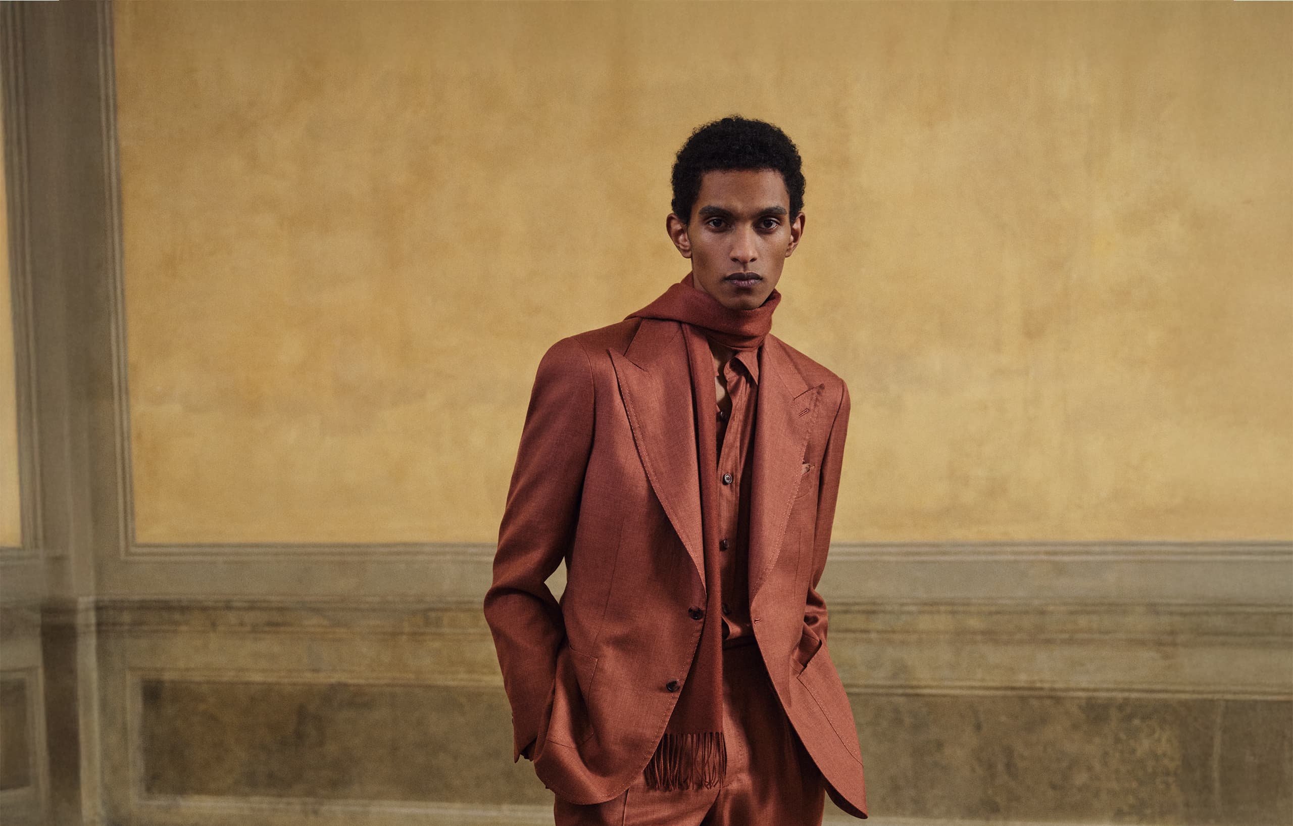 A Model wearing the Pompeian Red tailor suit of FW25 COLLECTION against a warm beige textured wall.