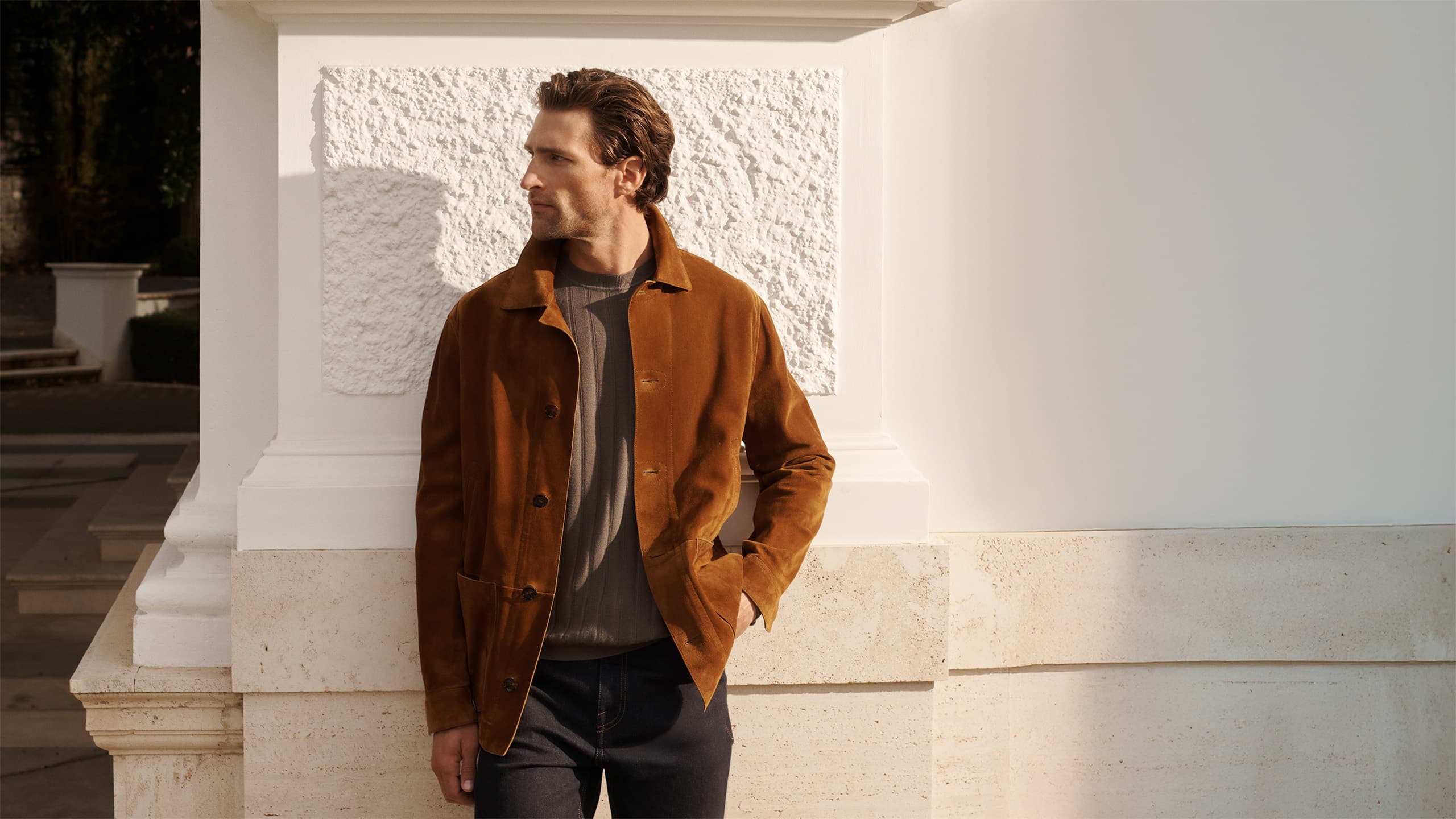 Man in Brioni rust-colored suede jacket and gray shirt standing against white textured wall in sunlight, looking to the side.