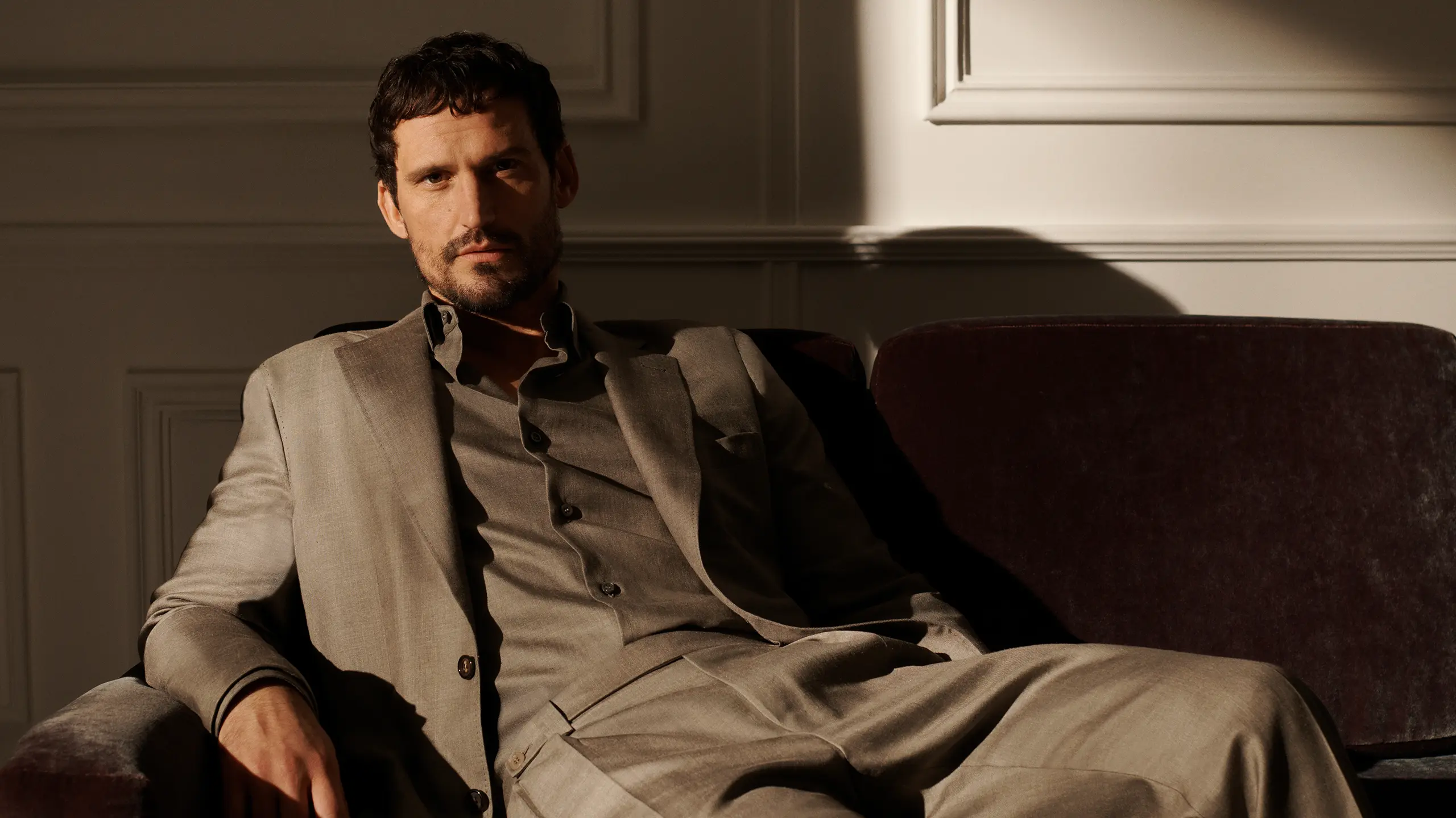 Man in Brioni beige suit sitting on brown velvet sofa in sunlit room with white paneled walls, looking directly at camera.