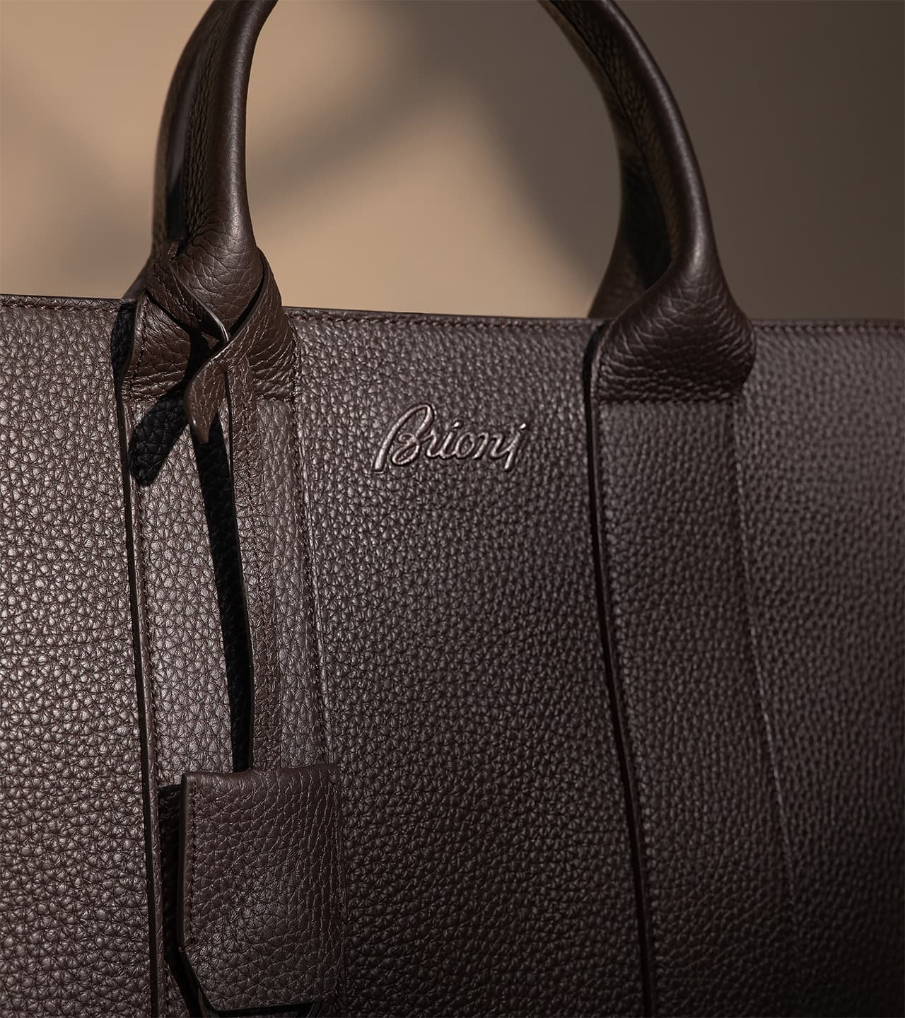 Close-up of a Brioni brown textured leather Brioni handbag with curved handles and embossed brand logo on the front.