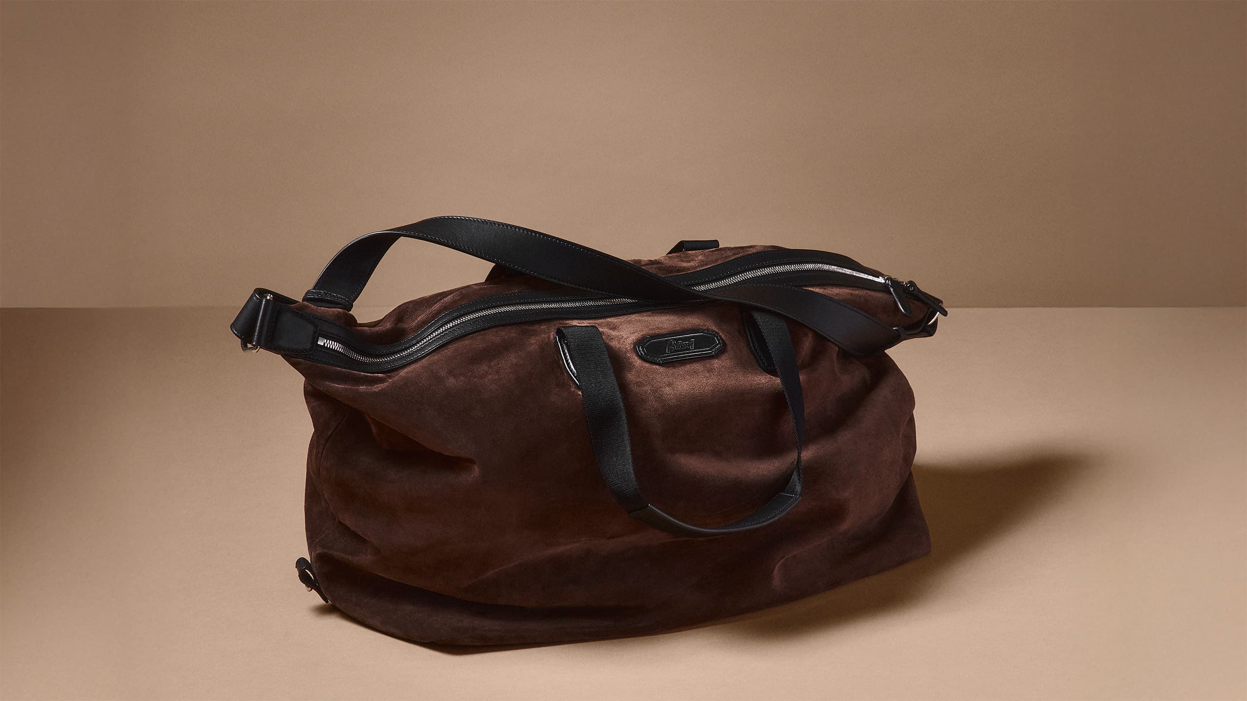 Brioni brown suede duffle bag with black leather straps and zipper details against a neutral beige background.