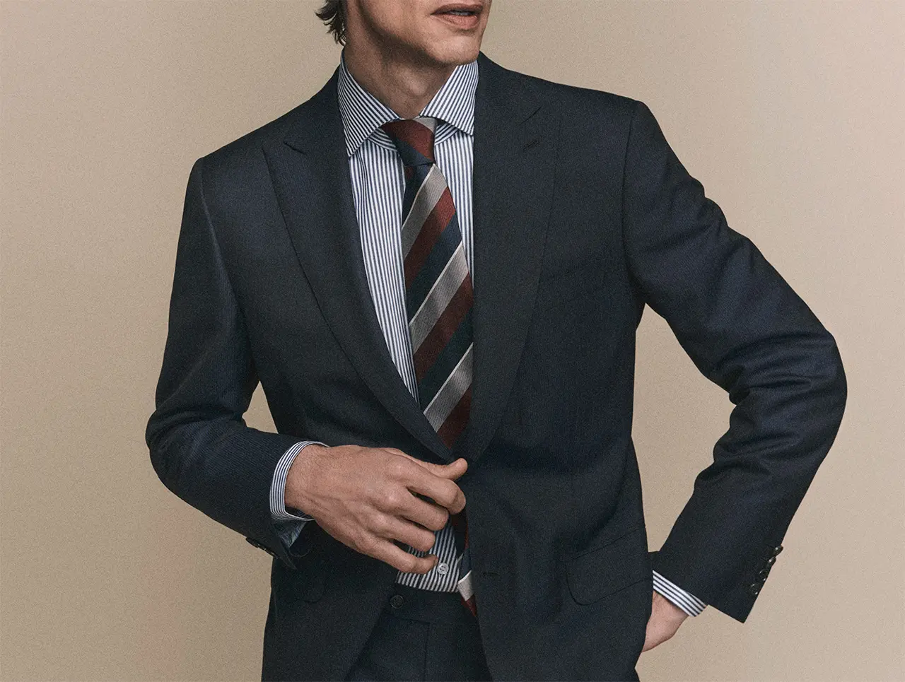 Person in Brioni navy suit with striped shirt and burgundy-gray striped tie against beige background.