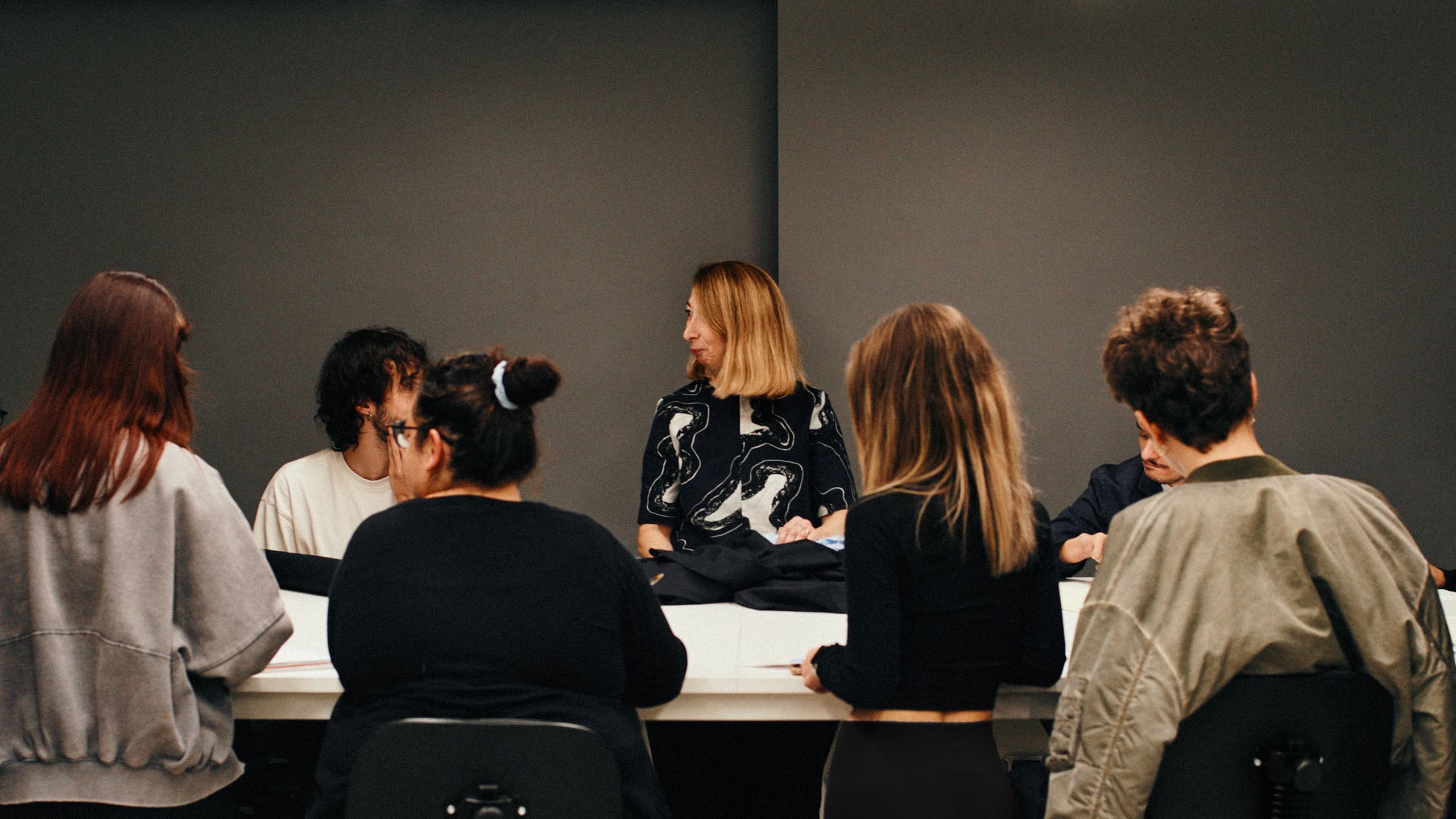 A Brioni tailor sits around table together with a group of students, showing them an item in the making.