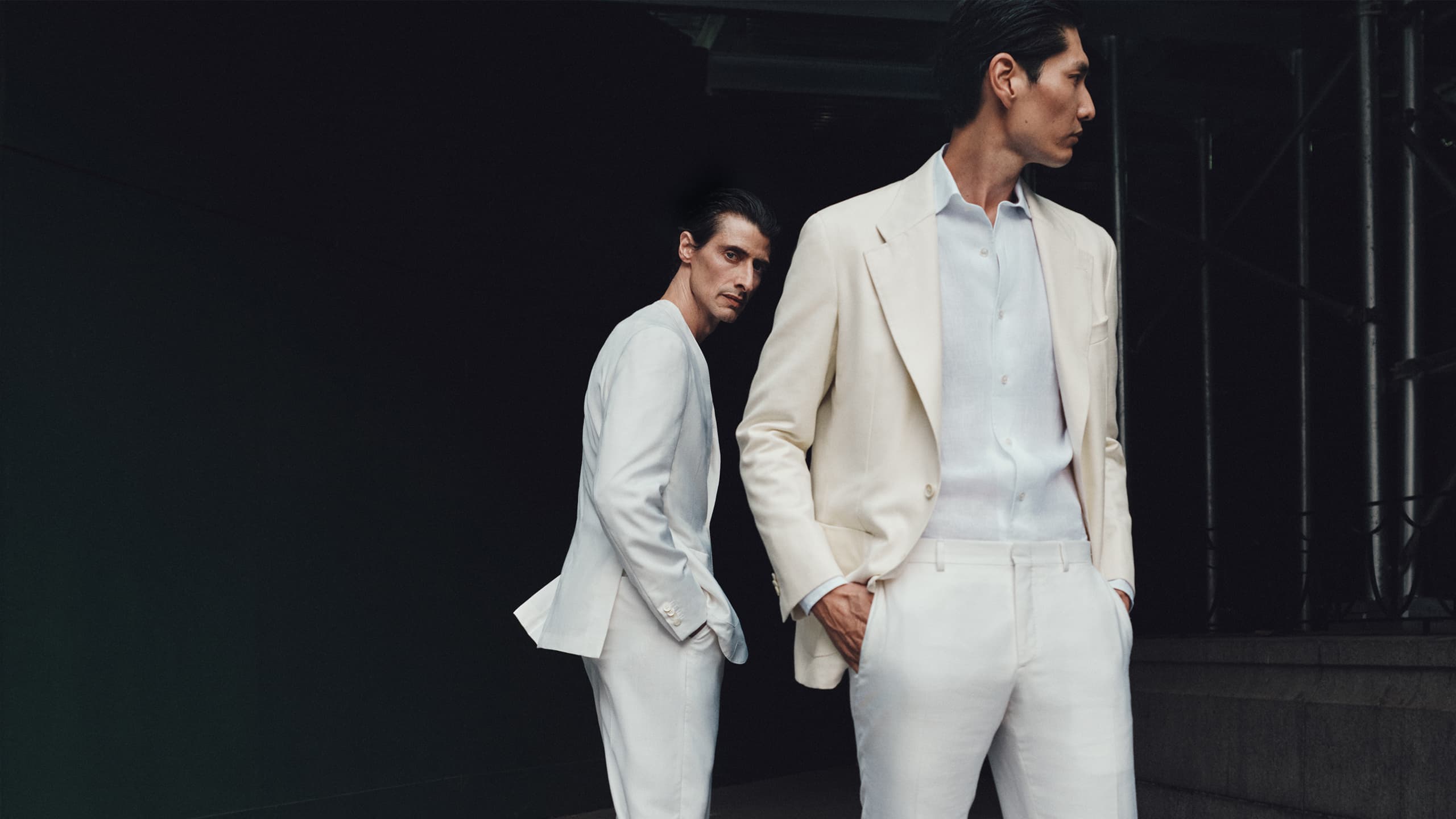 Two models in elegant Brioni white suits against a dark background, one in cream blazer and one in light suit.