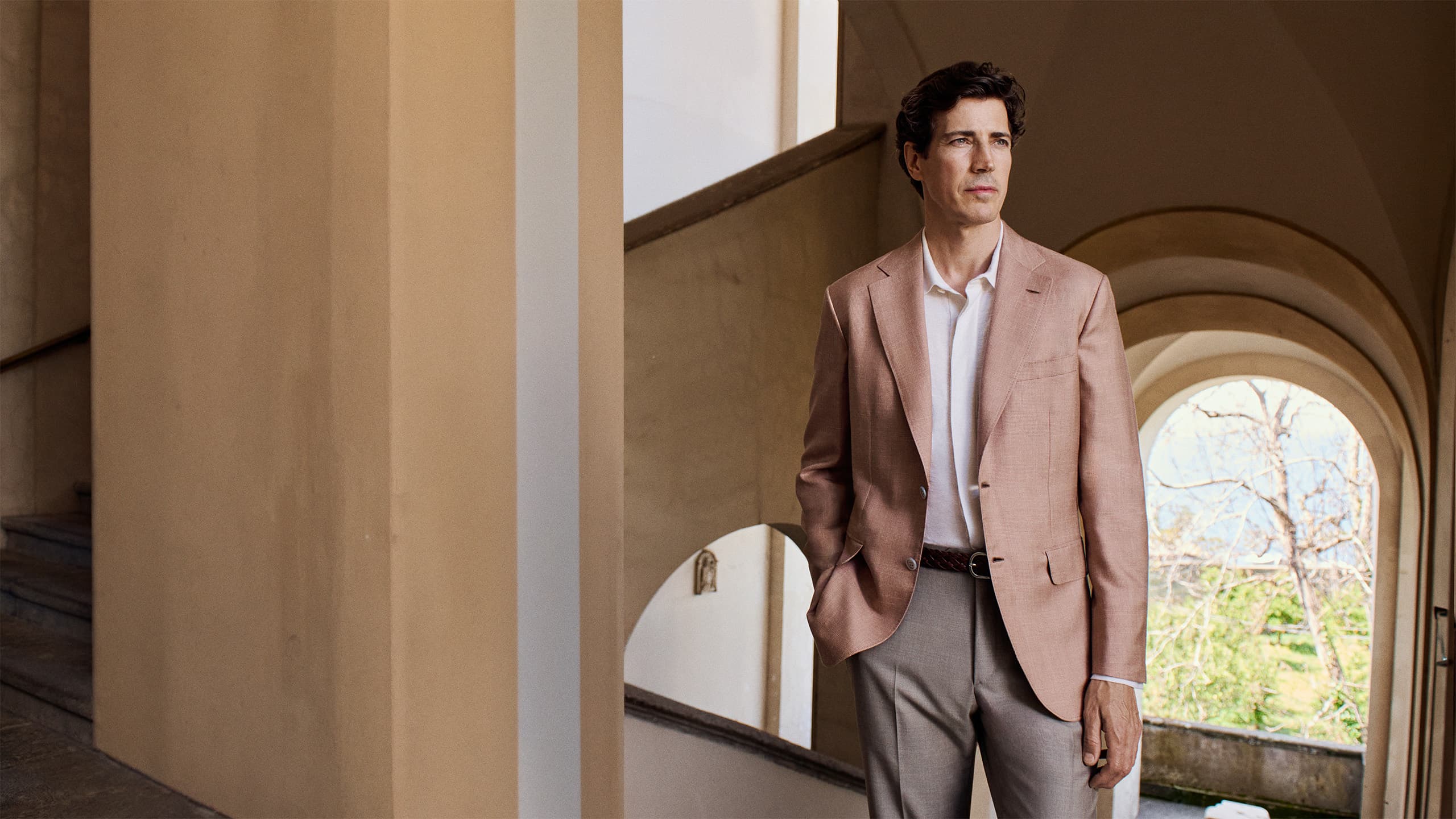 Man in Brioni beige blazer and white shirt standing in archway with warm neutral tones and natural light from arched windows.