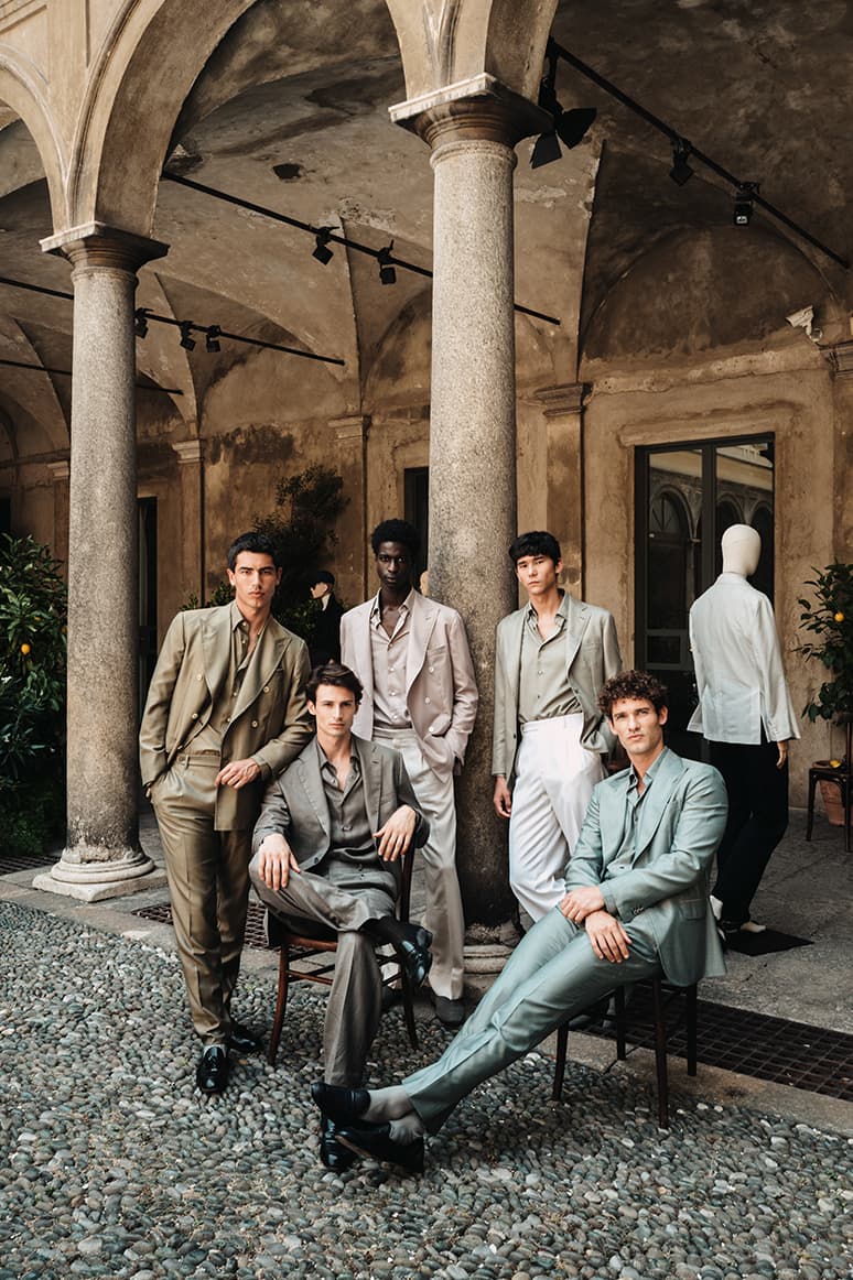 Models showing Brioni's ss26 collection in elegant neutral-toned suits posing in a historic Italian courtyard with stone columns and arched architecture.