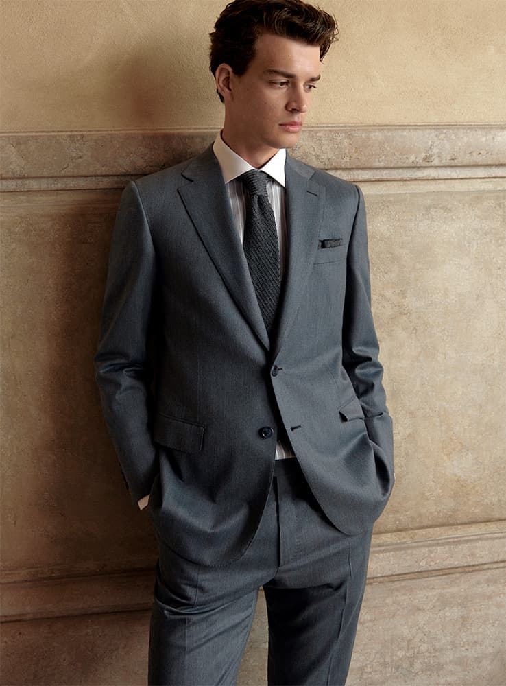 Young man in Brioni gray business suit with white shirt and dark tie standing against beige wall, looking sideways.