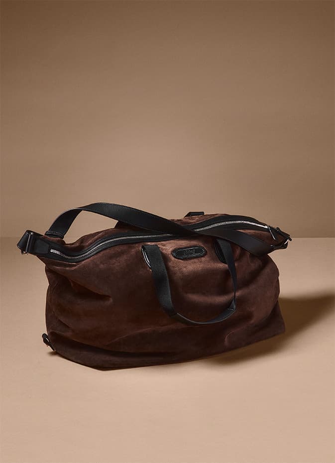 Brioni brown suede duffle bag with black leather straps against a neutral beige background.