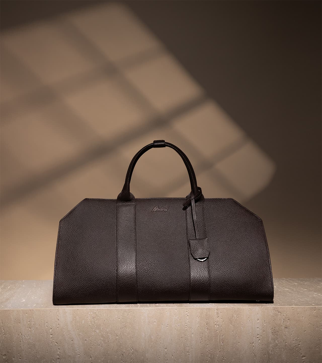 Brown leather Brioni handbag with handles and luggage tag on beige surface with geometric window light shadows.