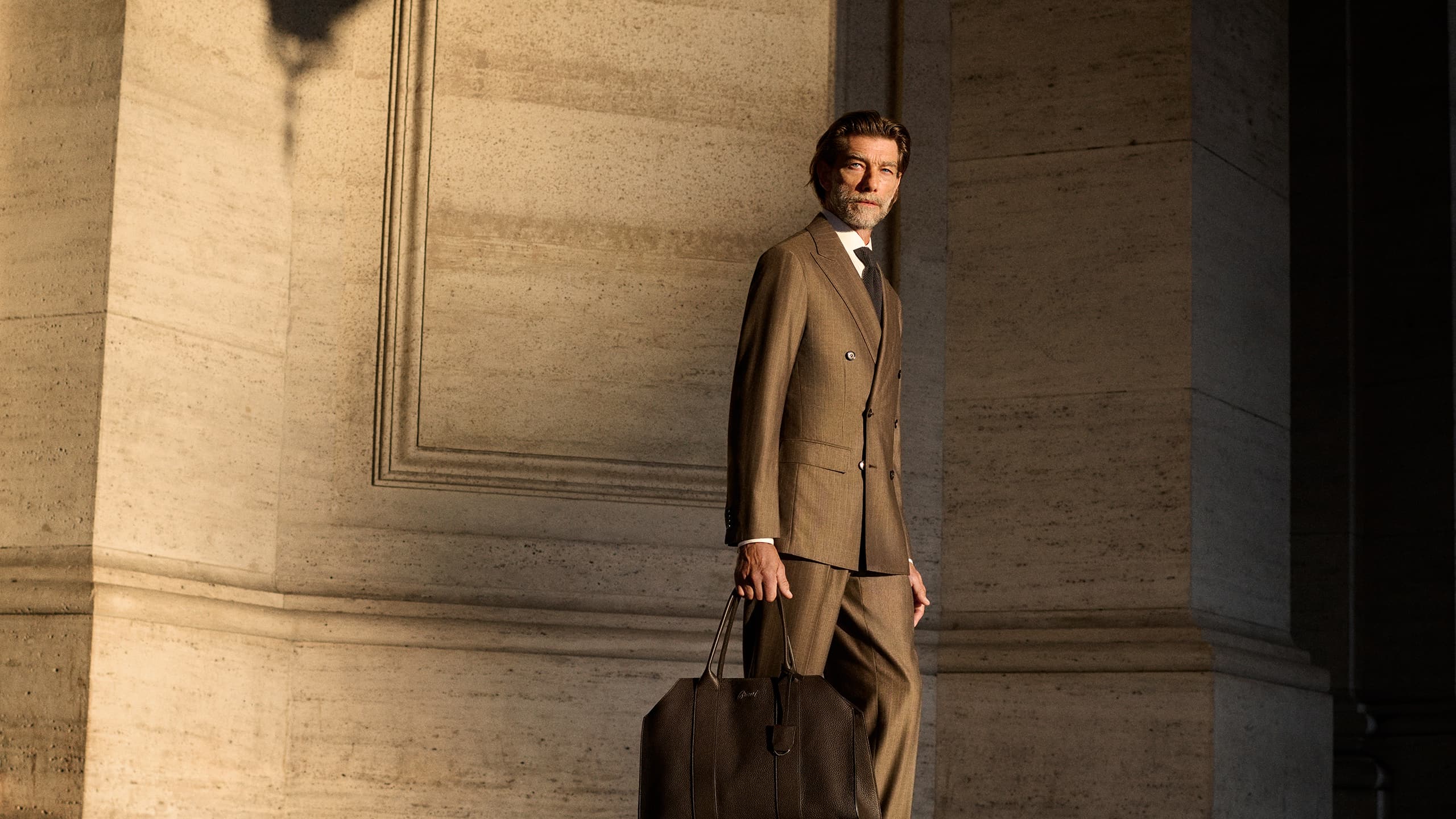 Man in Brioni tan suit holding brown leather bag stands against concrete wall in sunlight with dramatic shadows.