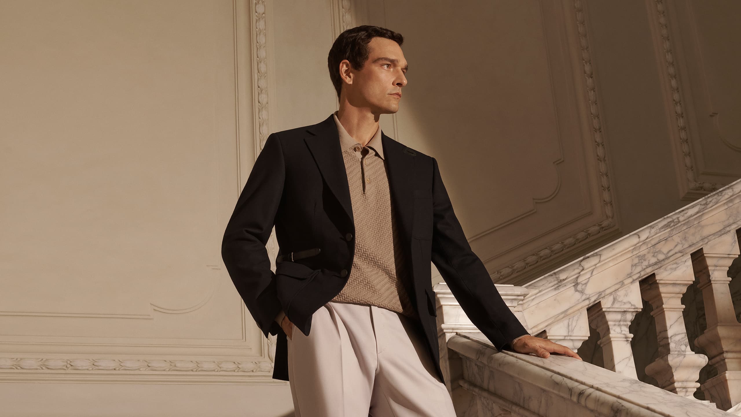 Person in black Brioni blazer and white pants standing beside marble balustrade in an elegant interior with ornate walls.