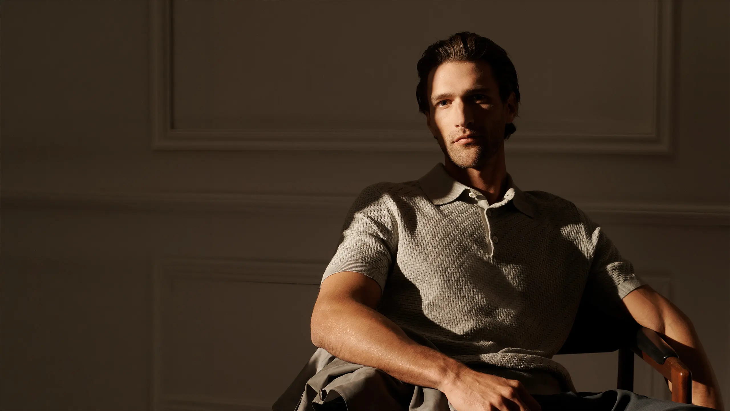Man with dark hair sitting in a chair wearing a Brioni gray and white patterned polo shirt against a neutral wall background.